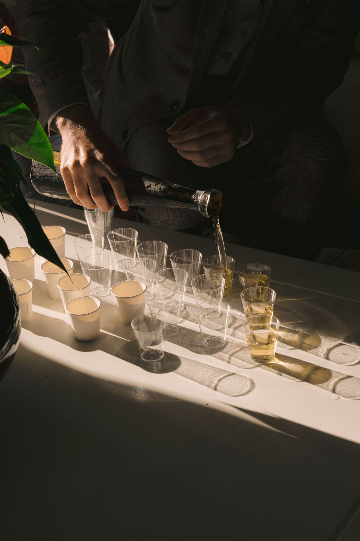Hands pouring drinks into multiple wine glasses arranged in a row on an illuminated white surface during a tasting event.
