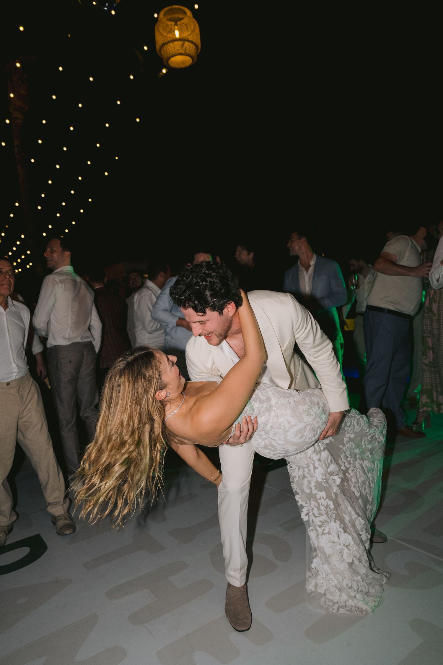 Couples dancing and dipping at a dimly lit wedding reception with lanterns overhead on a white dance floor.