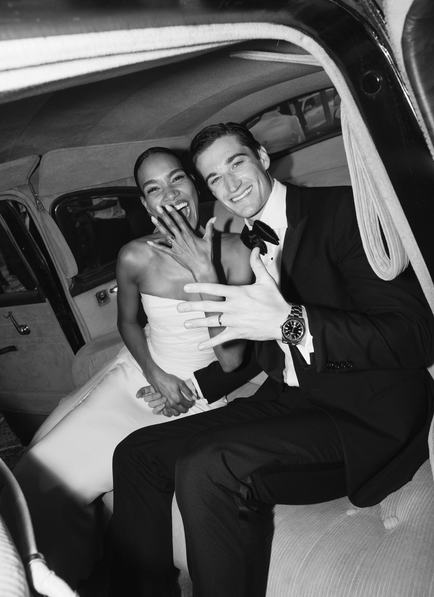 A well-dressed couple laughs joyfully while sitting in the back of a vintage car.