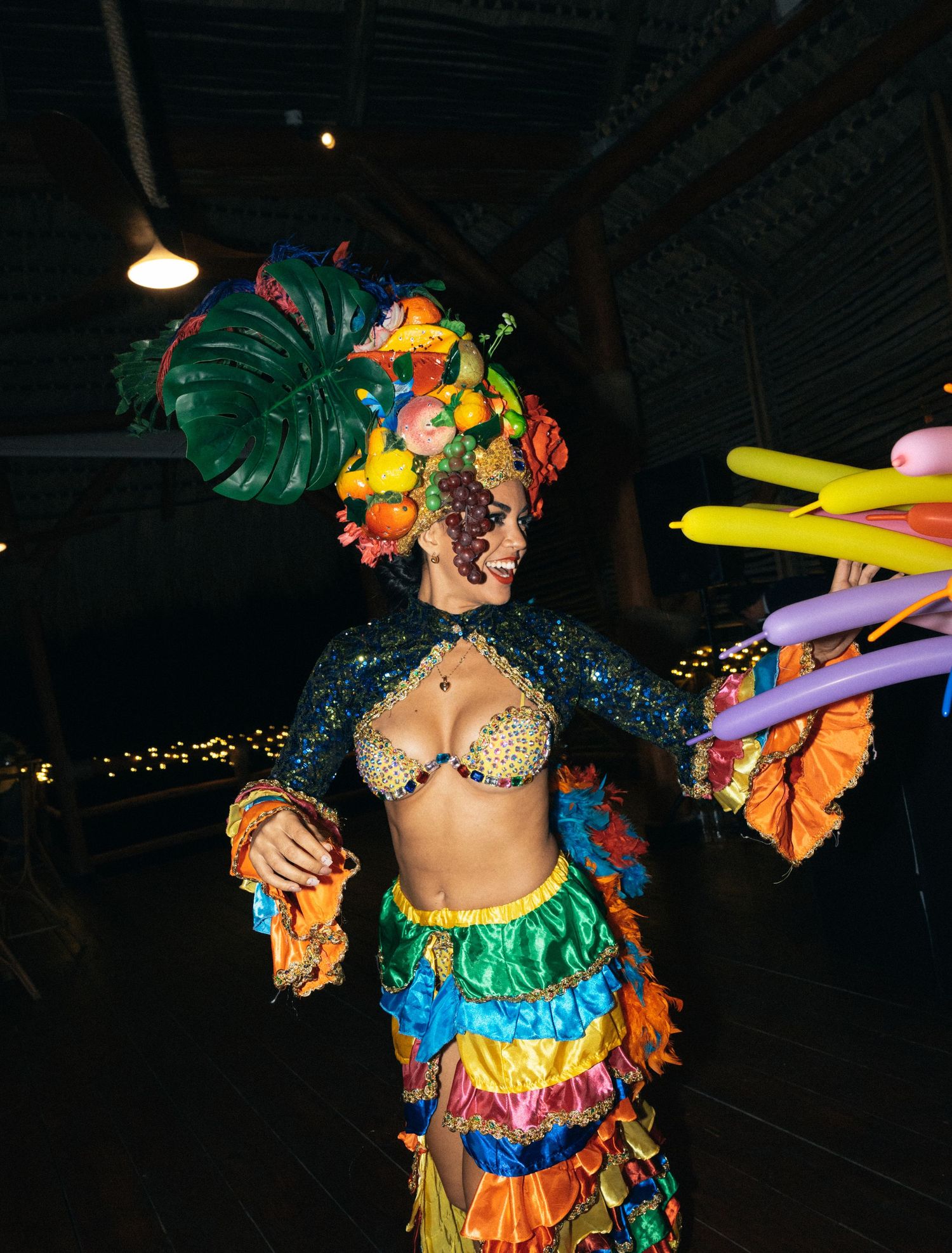Colorful festive carnival performer in tropical costume with monstera leaf headpiece and balloon decorations in dark venue.