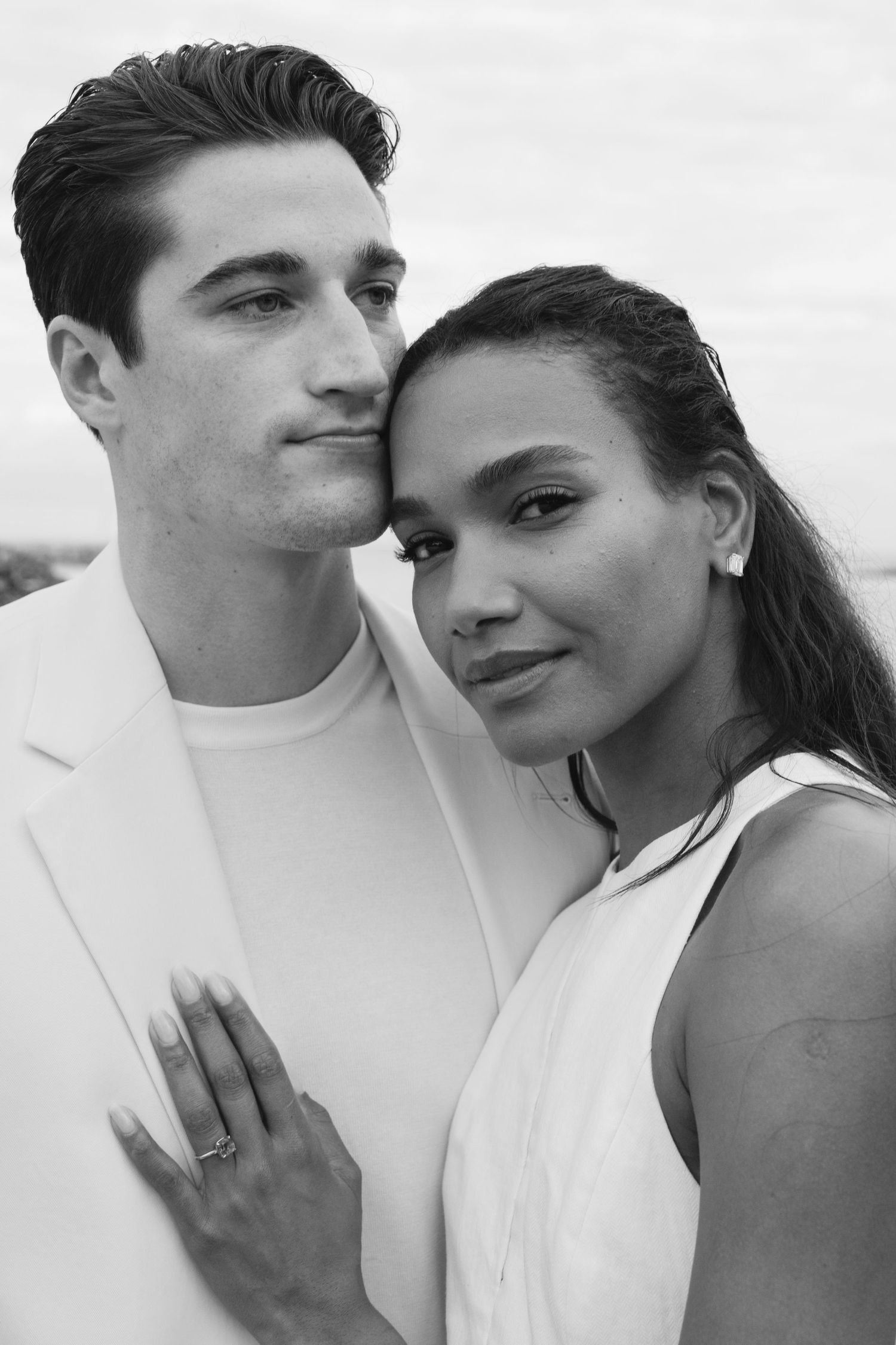 A romantic black and white portrait of a couple in white clothing embracing while looking at the camera.