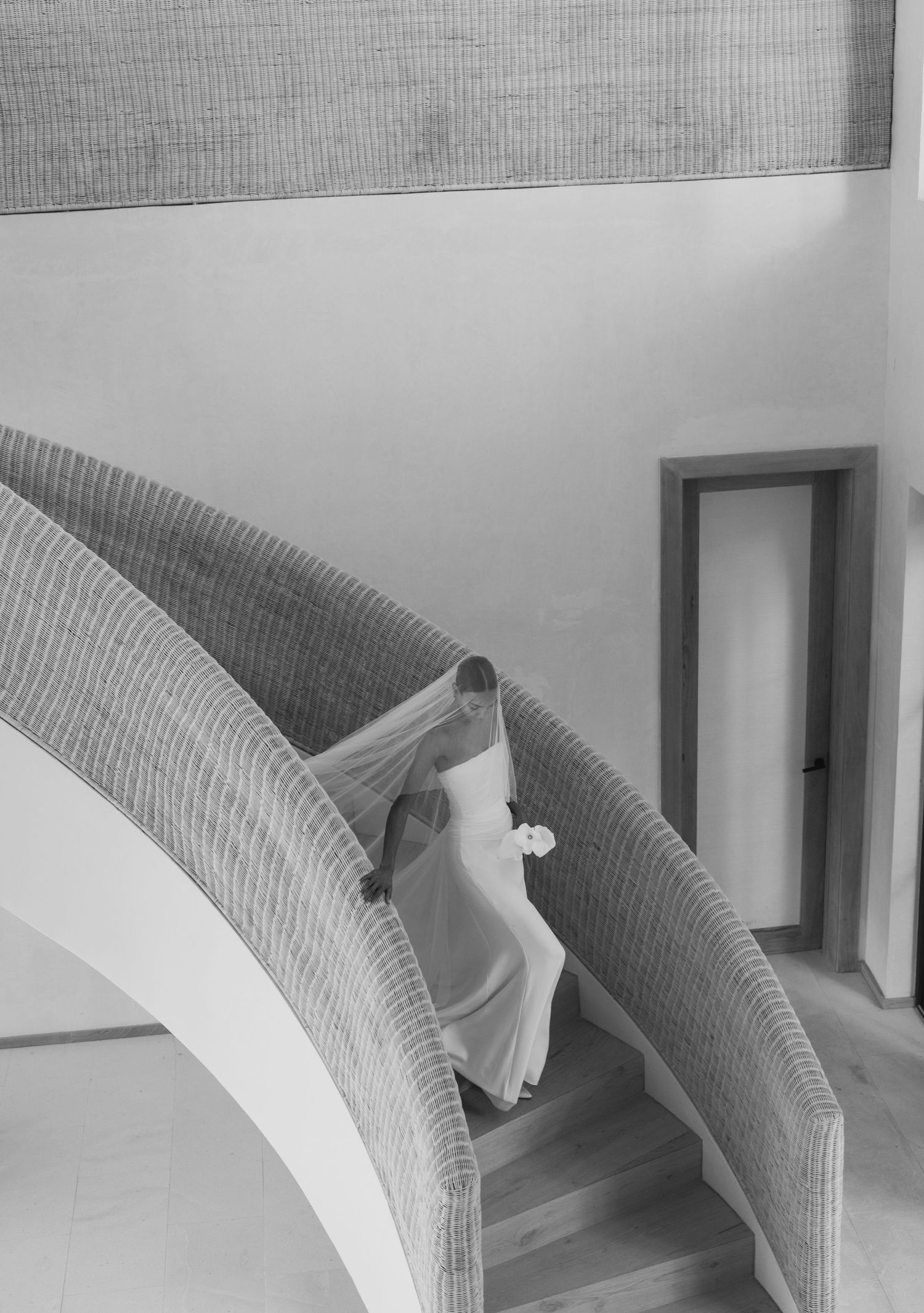 A figure in a flowing white dress ascends a curved staircase with textured walls in a black and white architectural image.
