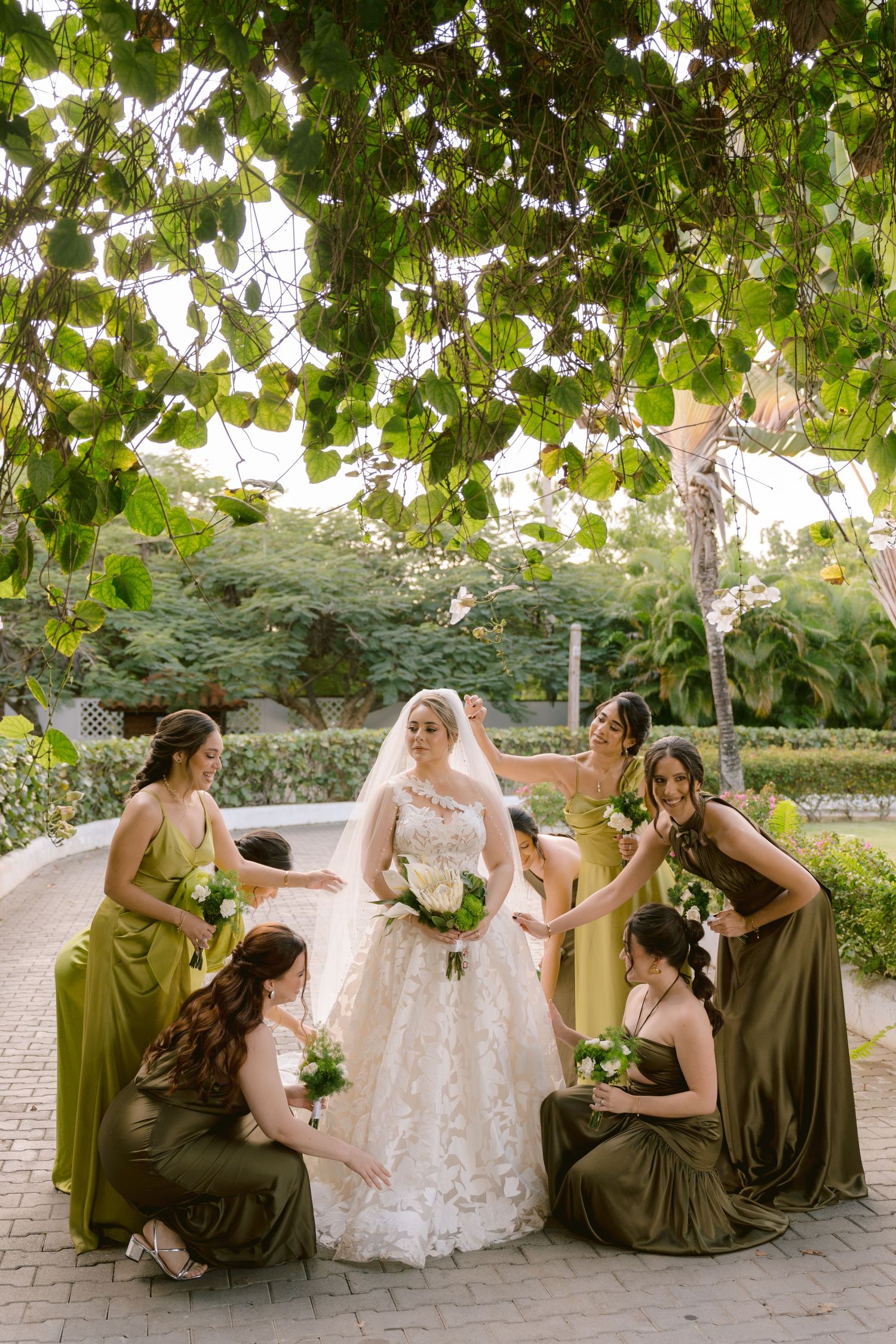 Wedding party in olive green bridesmaid dresses posing together in a garden setting.