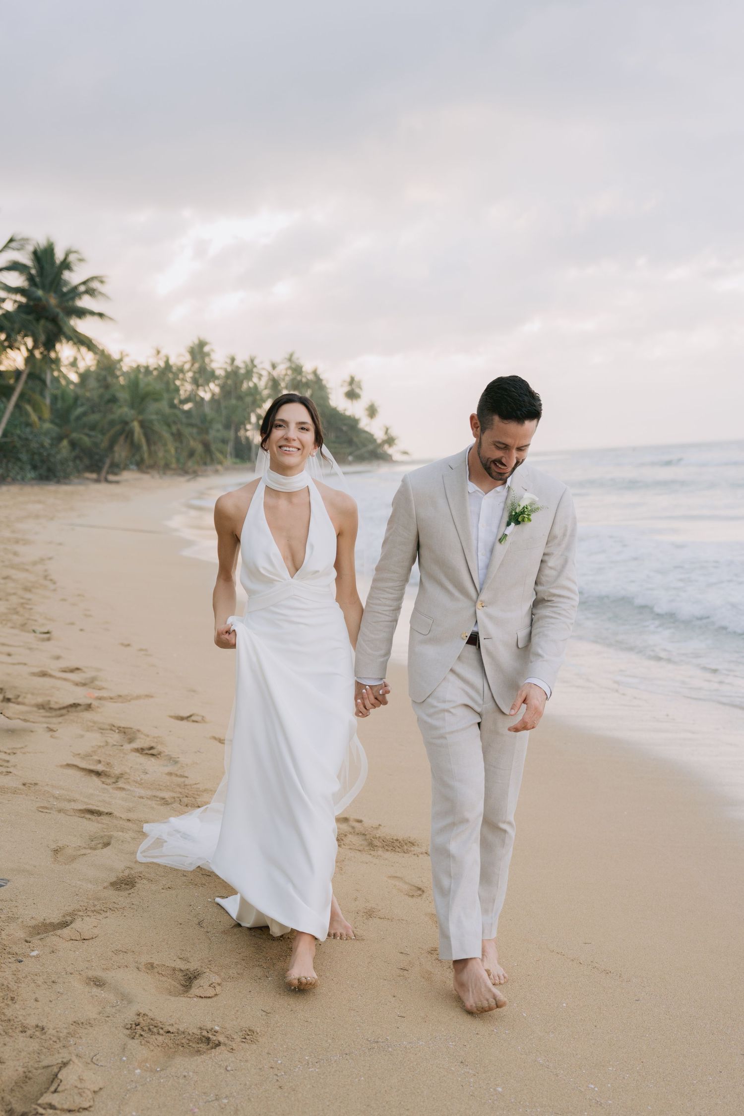 A newlywed couple walking barefoot along a tropical beach at sunset.