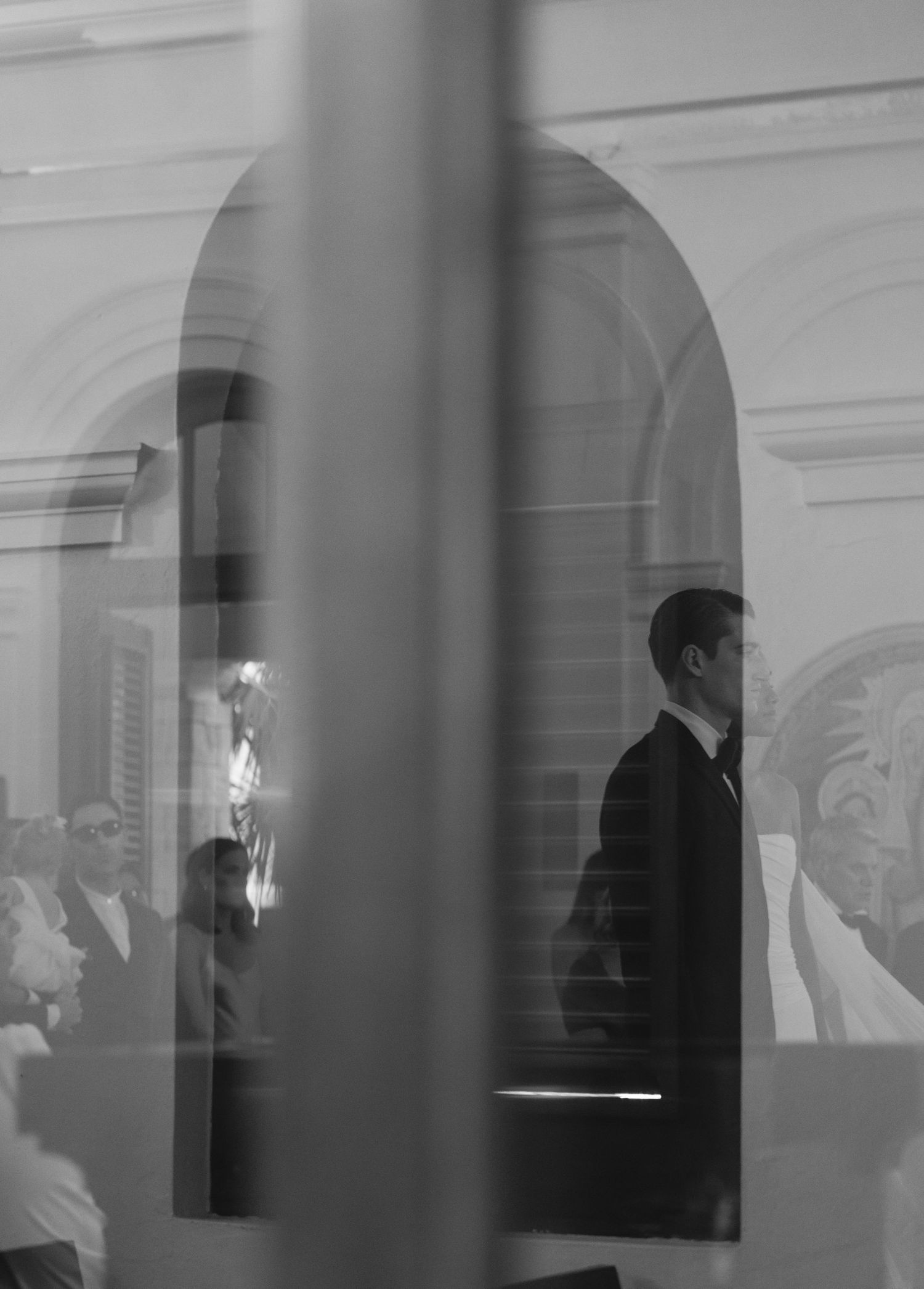 Black and white photo showing a figure in a suit viewed through architectural columns and windows in an elegant interior.