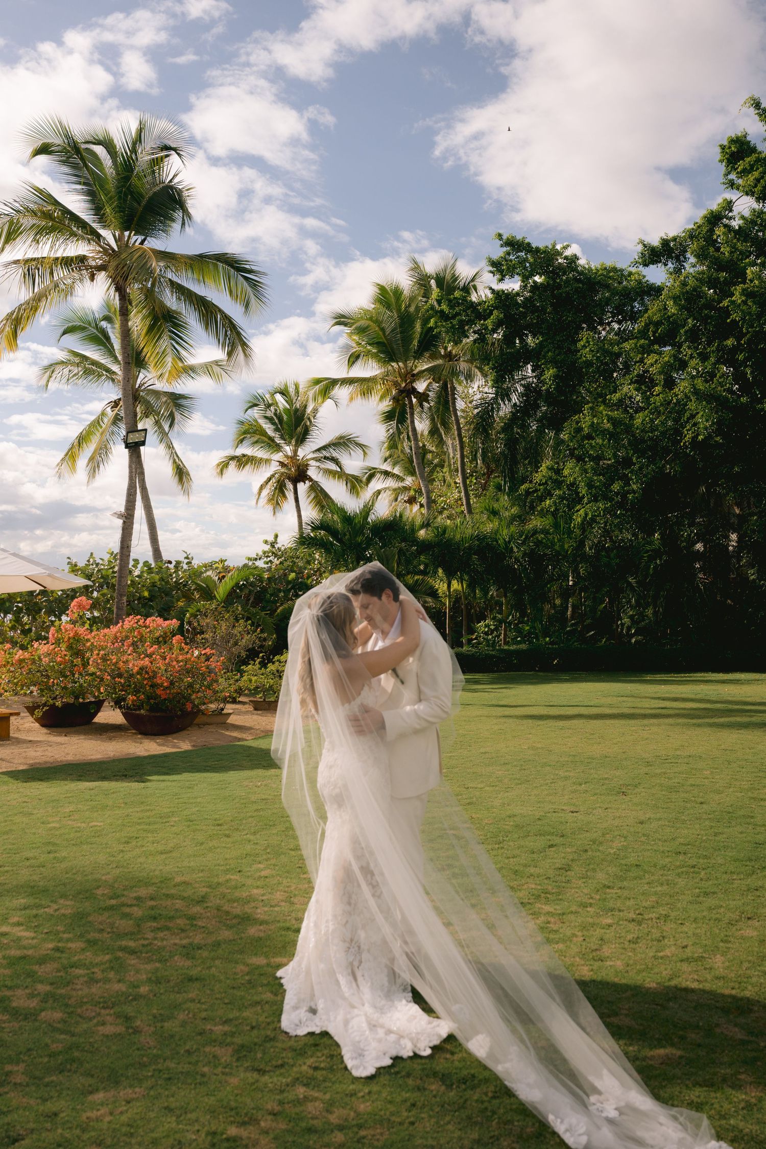 A flowing white wedding dress and veil dance in the breeze on a tropical lawn with palm trees and vibrant flowers.