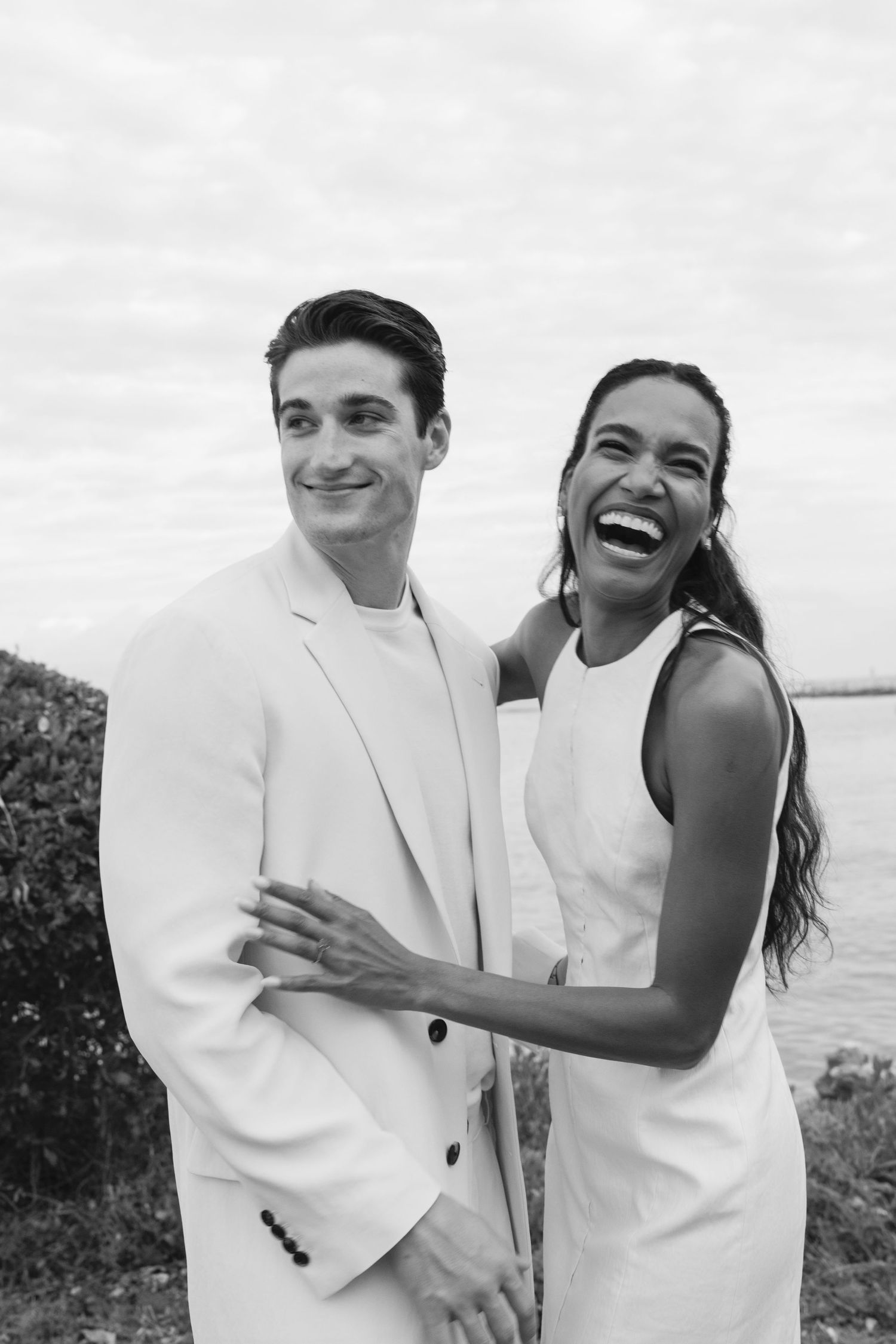 A happy couple dressed in white elegant clothing share a candid moment together outdoors by the water.