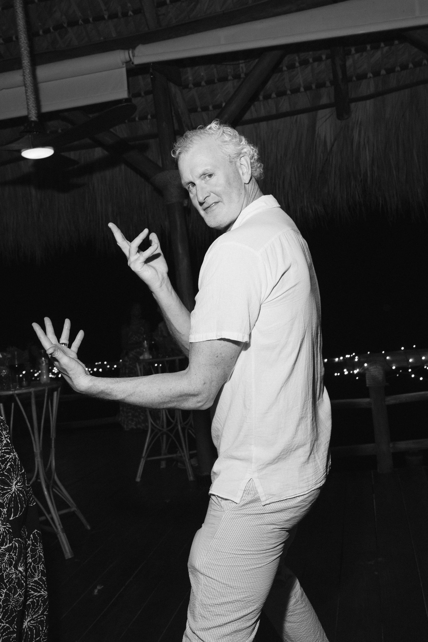 A person in a white t-shirt and light colored pants gestures while standing in a dimly lit venue at night.