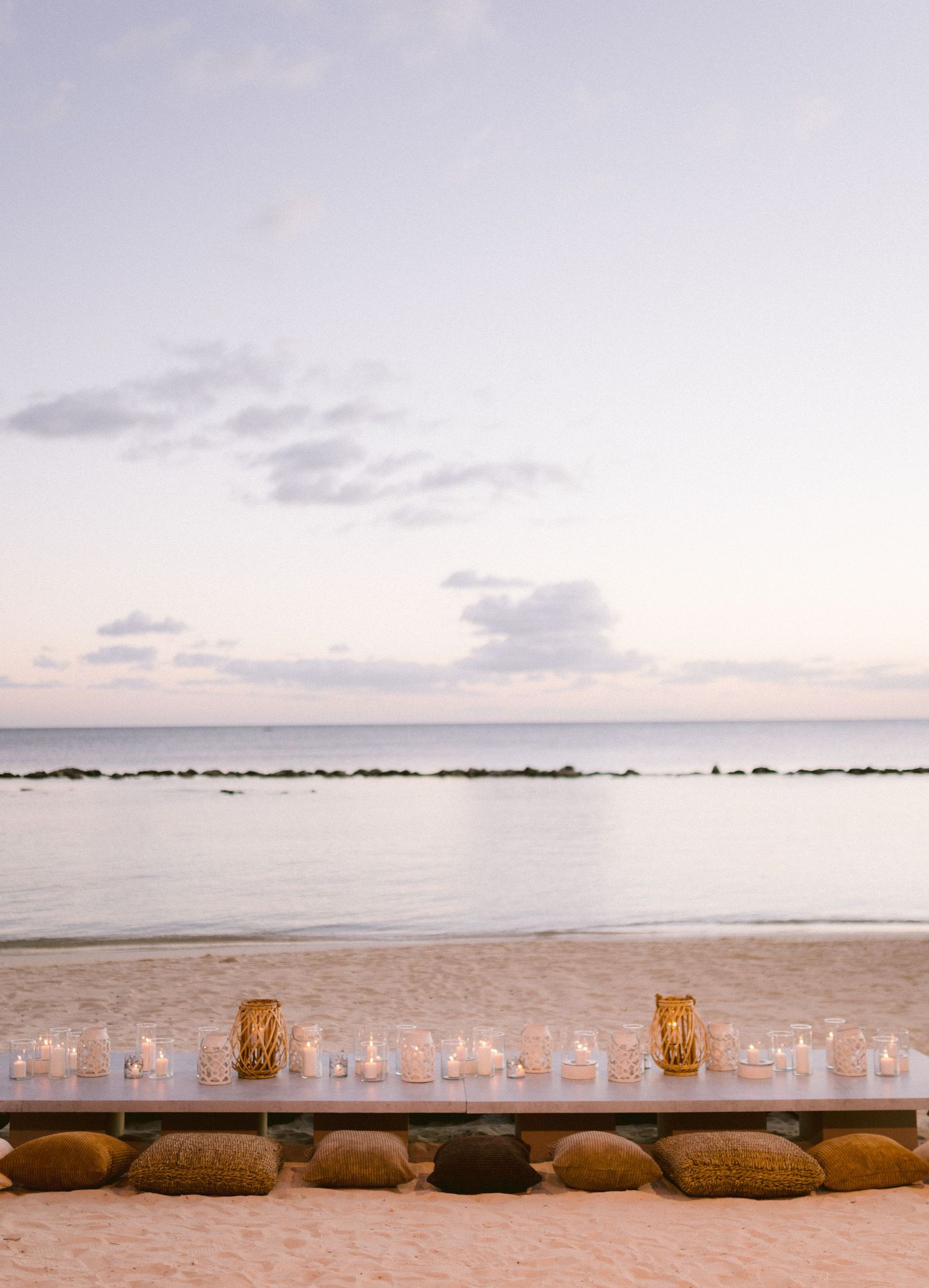 Romantic beach setup with candles in glass holders and floor cushions at sunset.