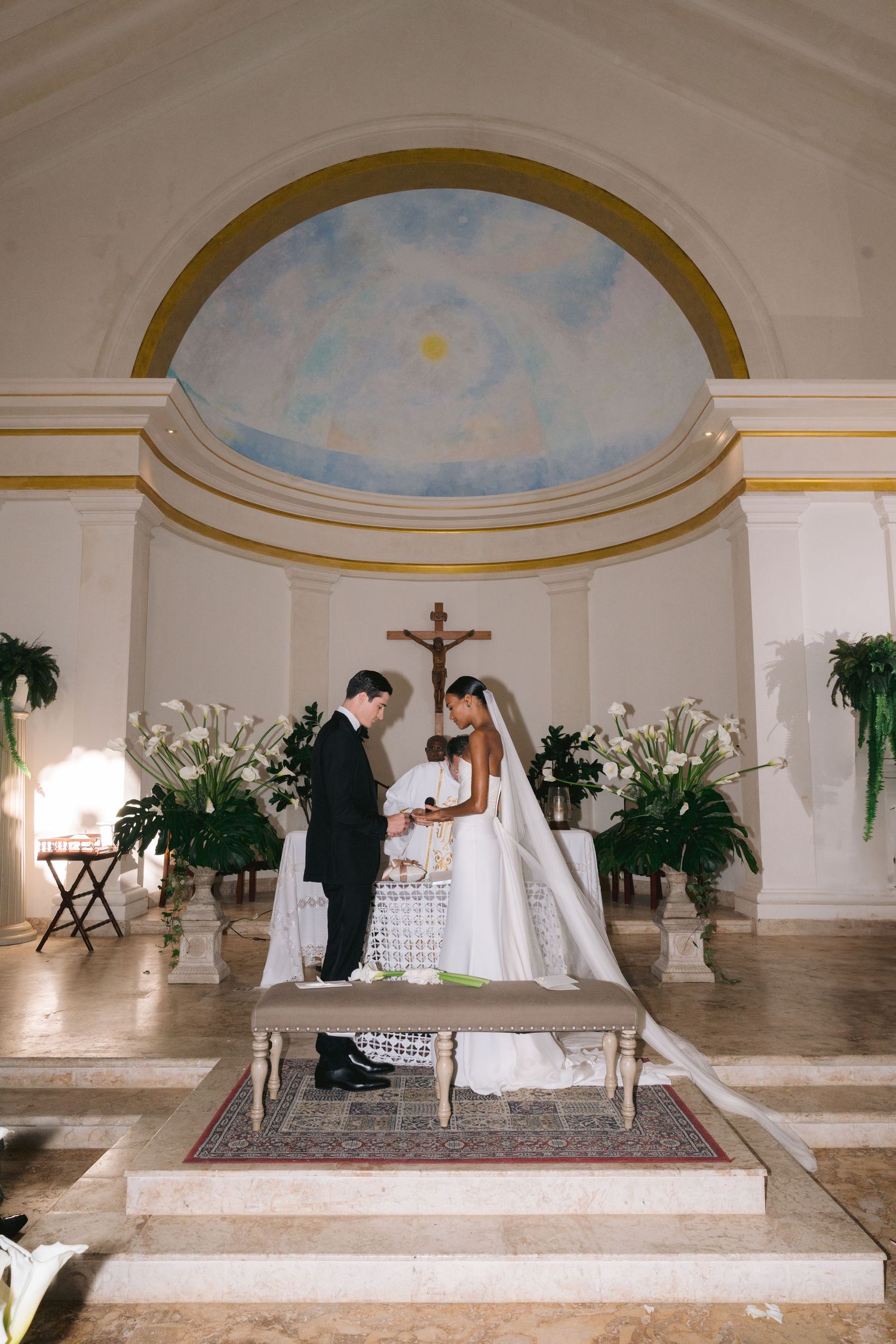 A wedding ceremony takes place in an elegant church chapel with a painted sky dome ceiling and white lily floral arrangements.