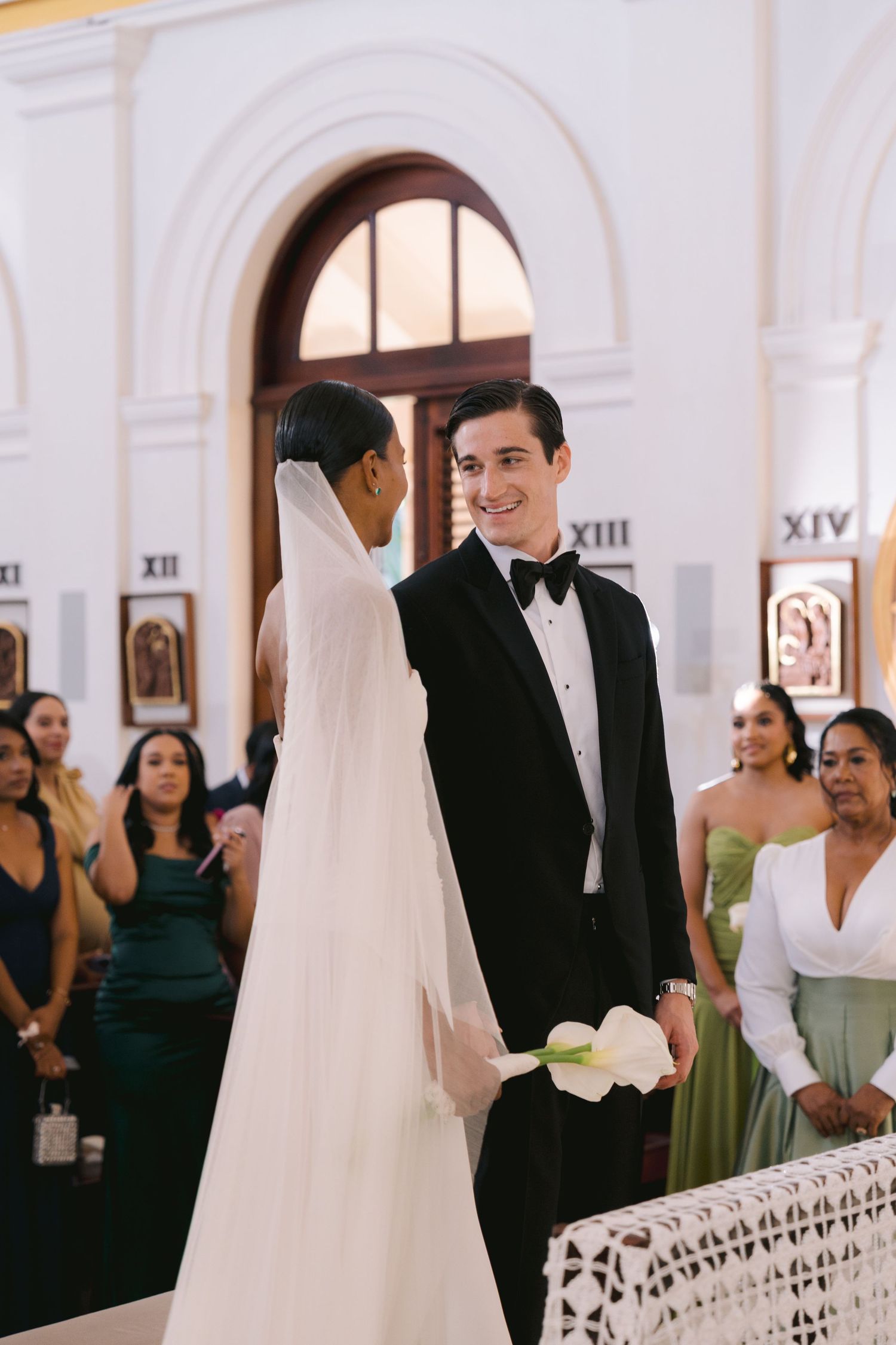 A wedding ceremony takes place in a church with arched doorways and white walls as guests look on.