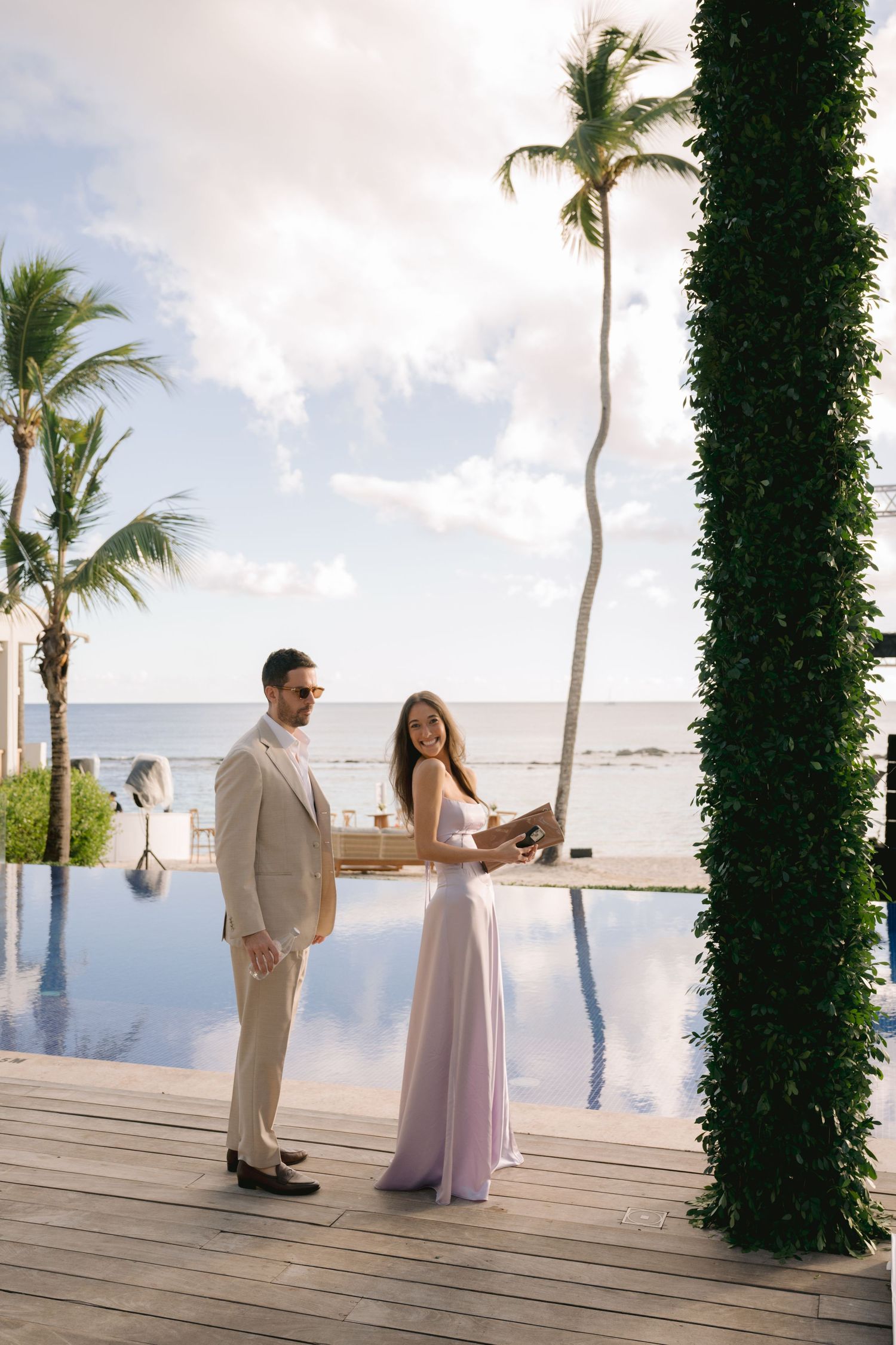A couple stands on a wooden deck by an infinity pool overlooking the ocean with palm trees and tropical scenery.