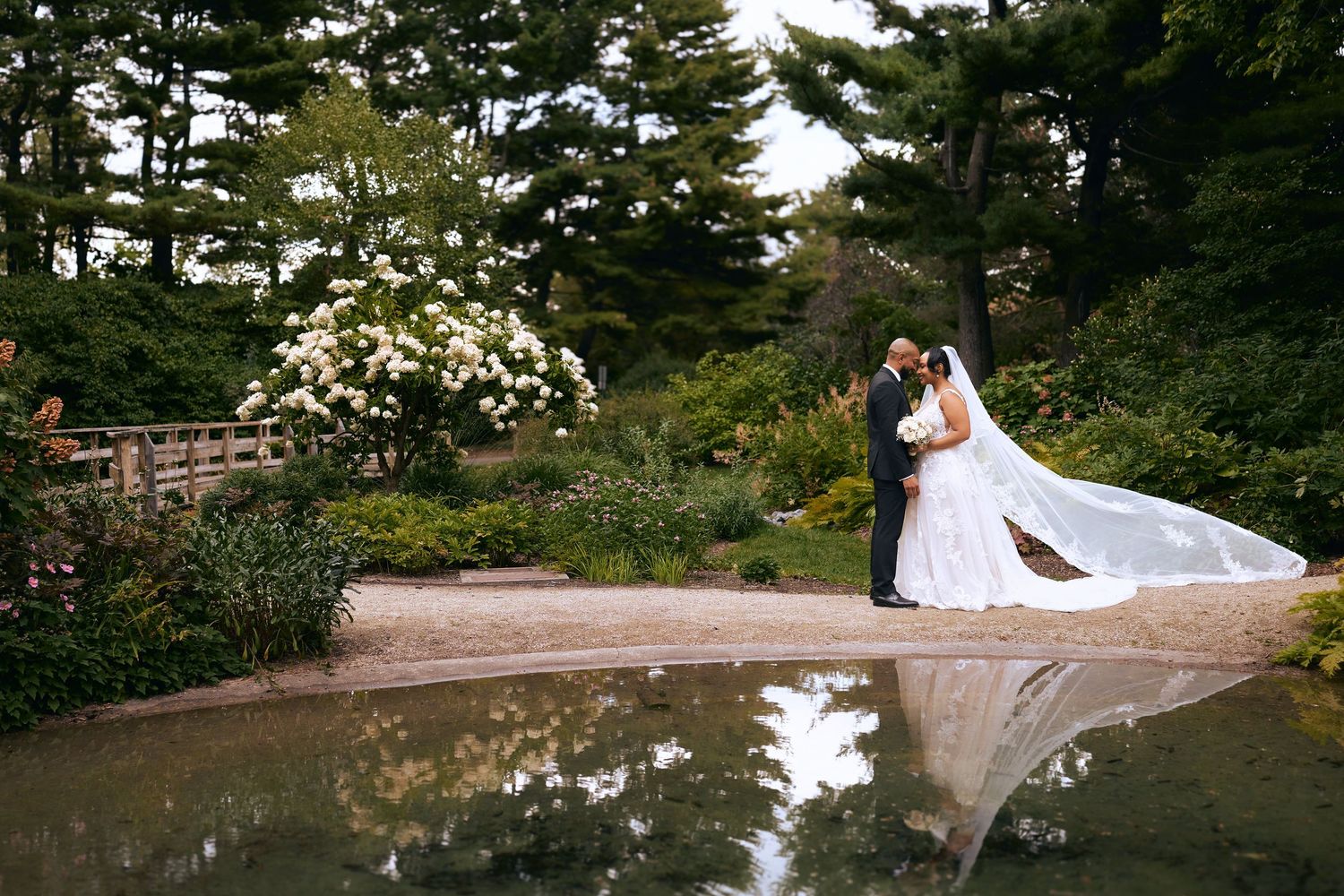 Bride and groom share a kiss beside a reflective pond surrounded by lush garden greenery at outdoor wedding.