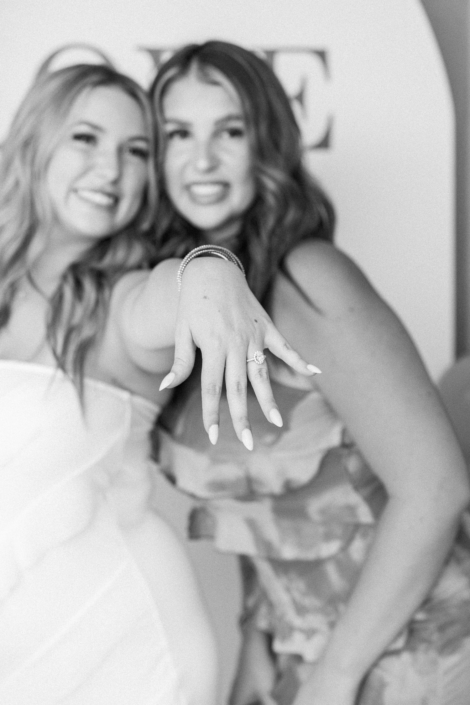 woman holding hand out in front of her showing off her engagement ring next to friend in black and white