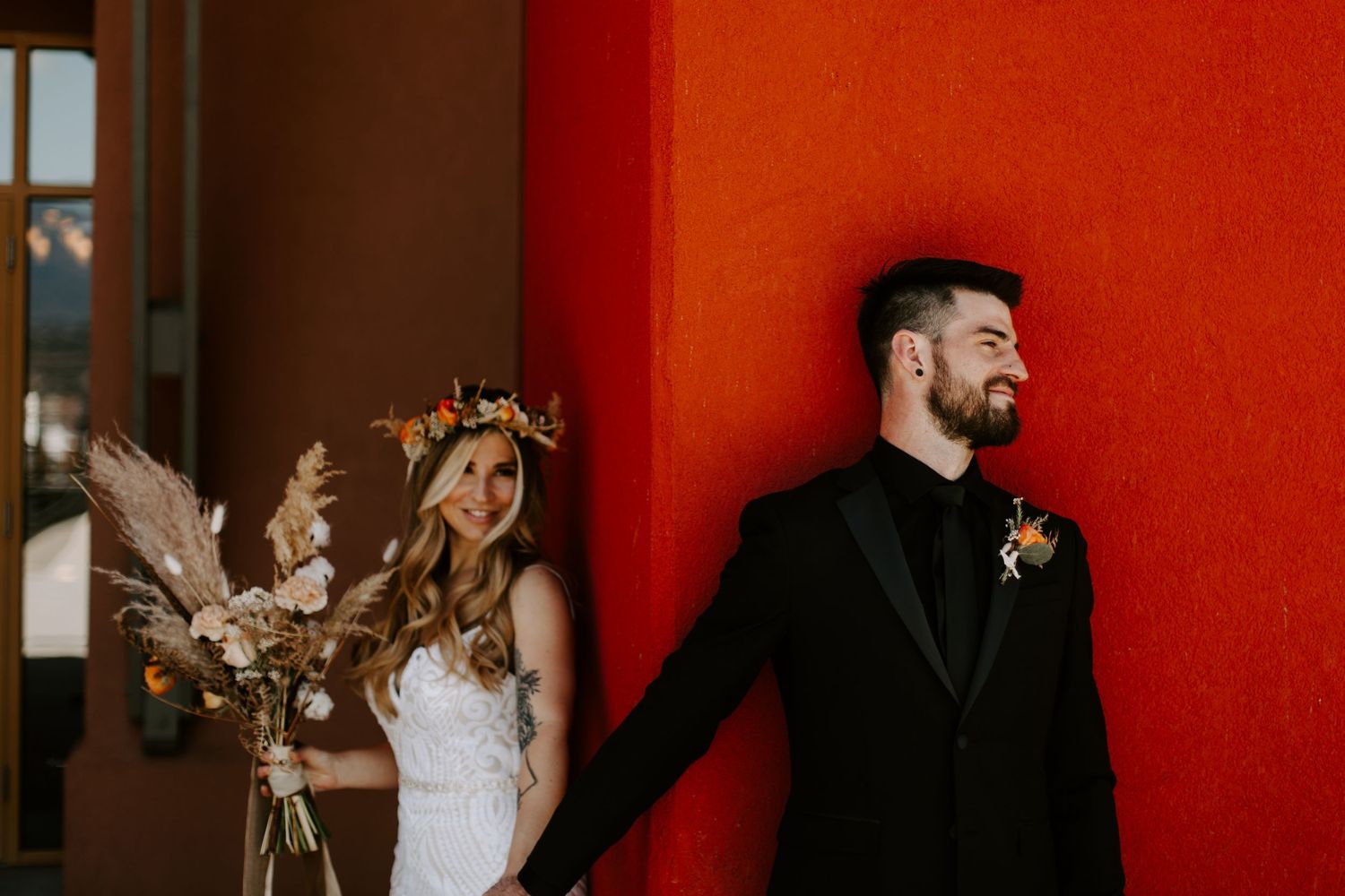 Wedding couple poses against vibrant red wall with dried floral arrangements.