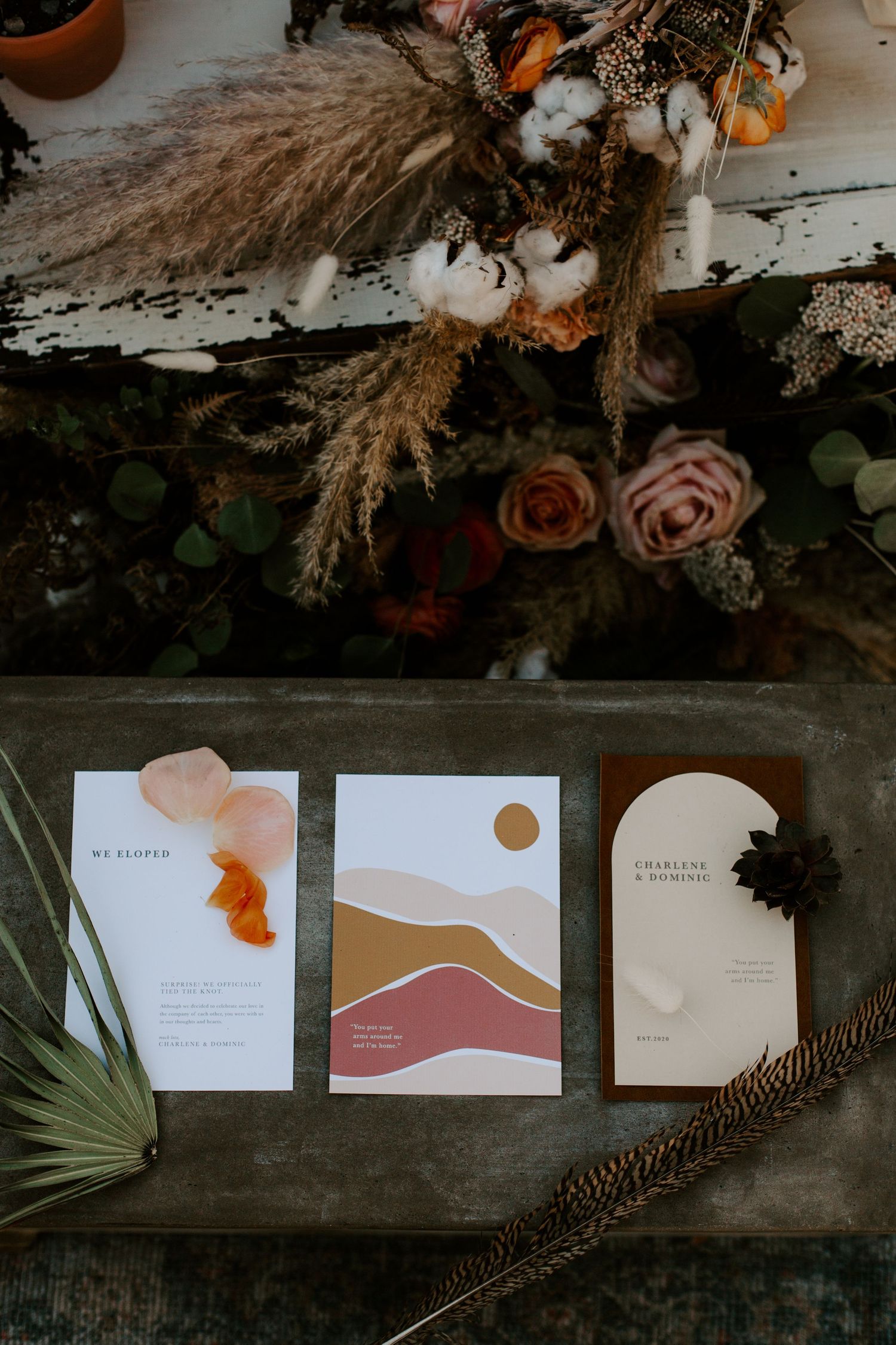 Modern minimalist wedding stationery with abstract organic shapes and dried floral decorations on a dark wooden surface.