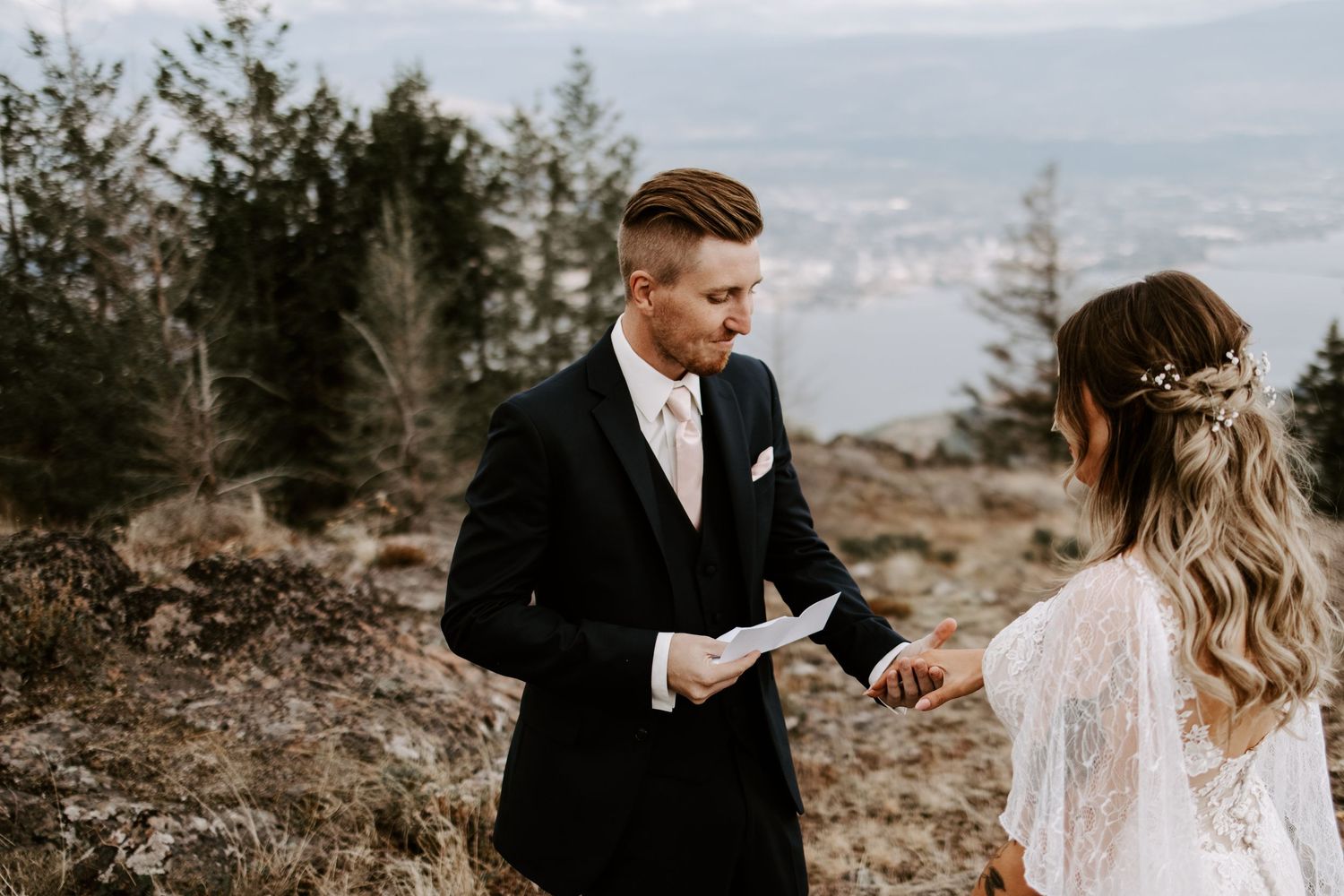 A couple exchanges vows on a scenic mountaintop with lace dress details and formal attire.