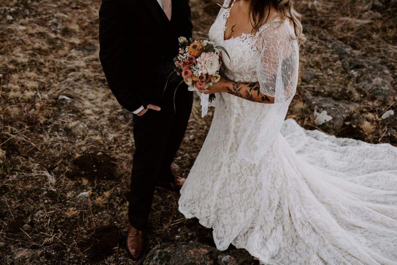 A couple stands on rocky terrain with the bride holding a colorful bouquet in her lace gown.
