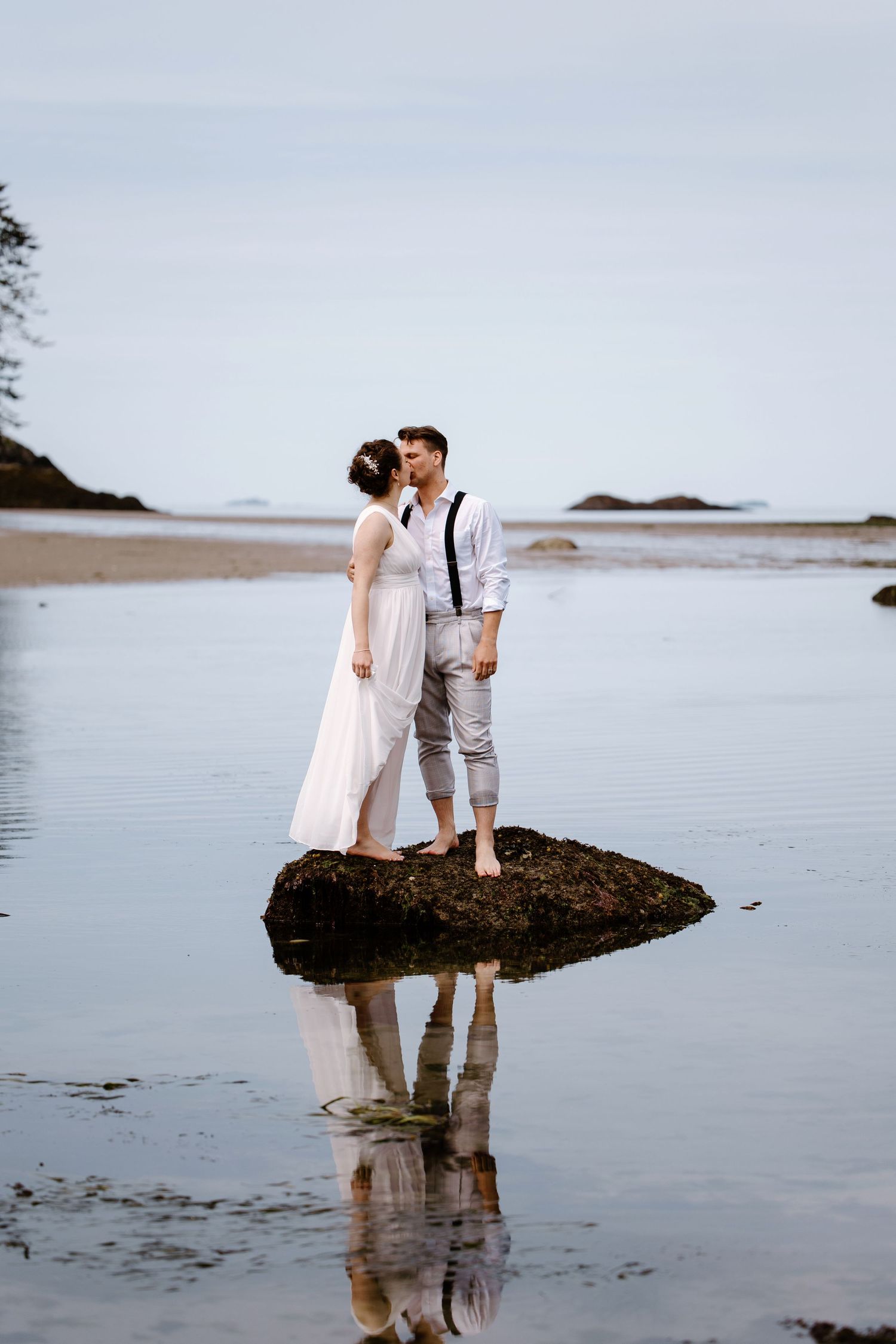A couple in wedding attire share a romantic moment while standing on a rock at the beach with their reflection in the water.