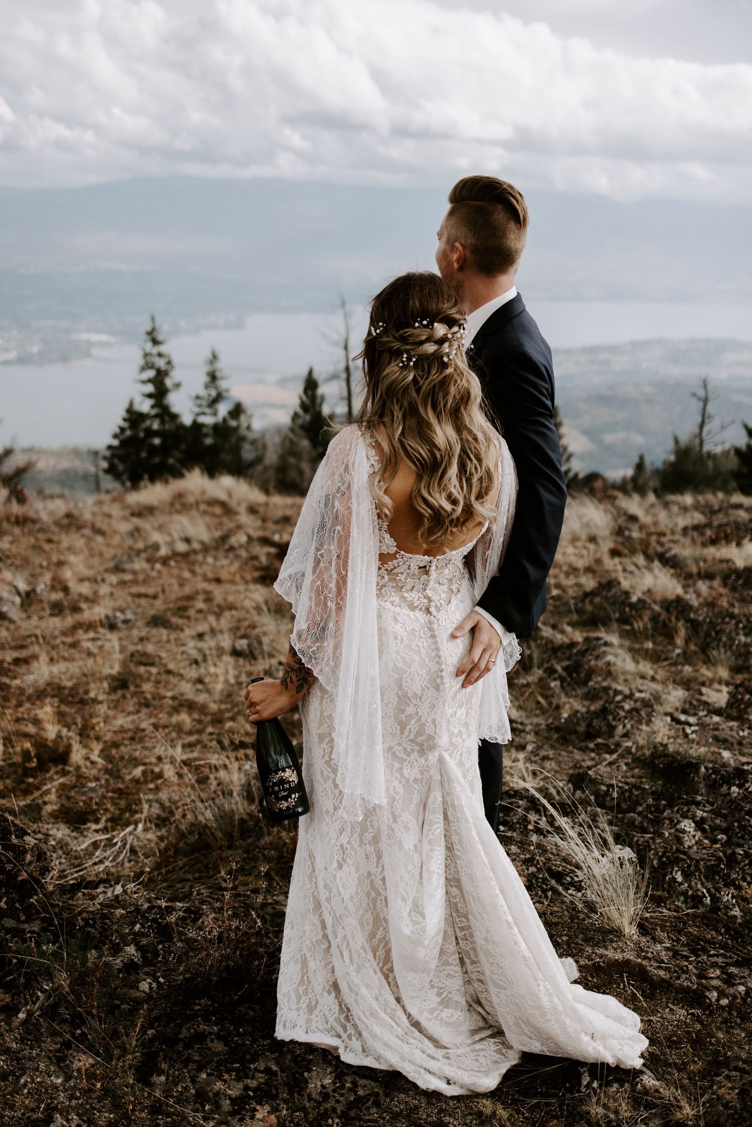 A couple in wedding attire embraces on a scenic mountaintop with dramatic cloudy skies in the background.