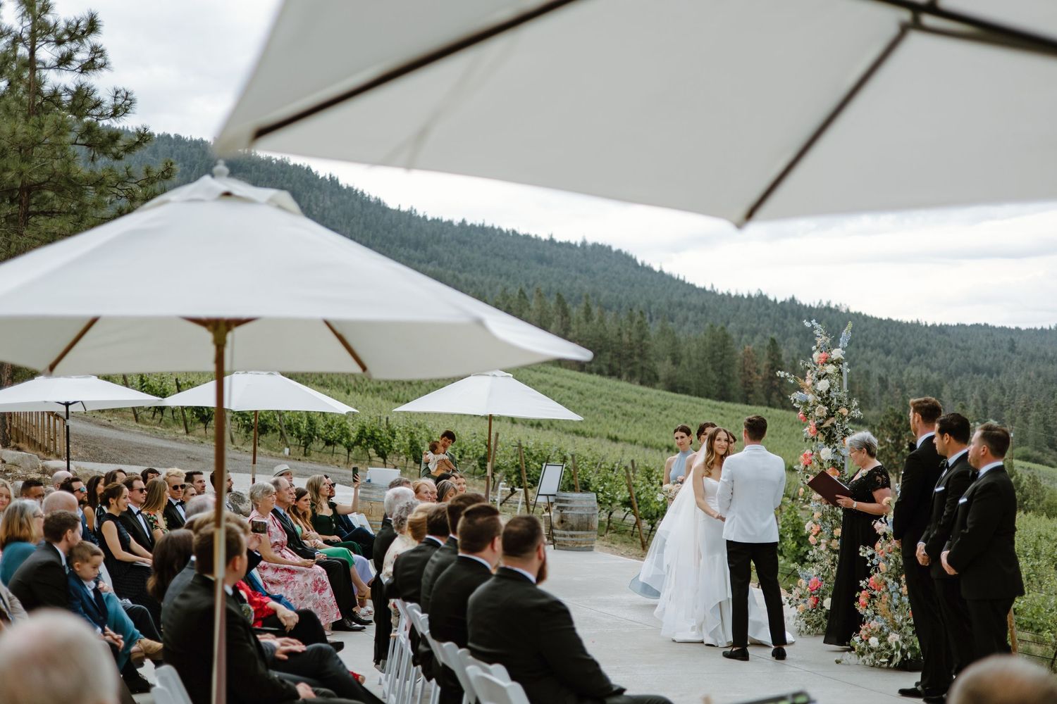 Outdoor vineyard wedding ceremony with guests seated under white umbrellas, couple at floral altar, forested hills backdrop.