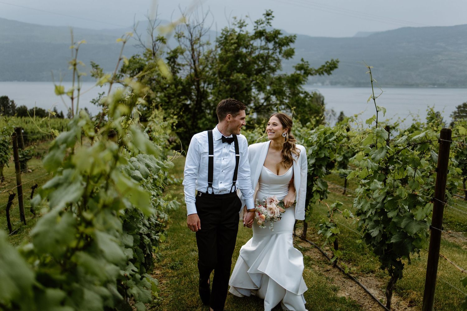 Bride and groom walk hand in hand through lush vineyard rows with scenic lake views on their wedding day.