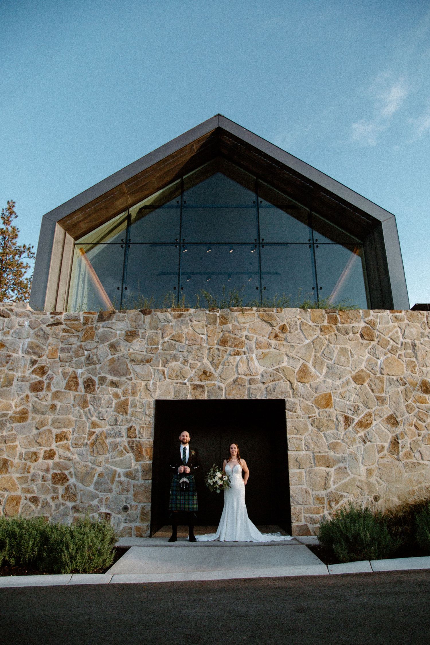 Bride and groom stand in stone building doorway with large glass gable window, groom wearing tartan kilt, bride in white gown.