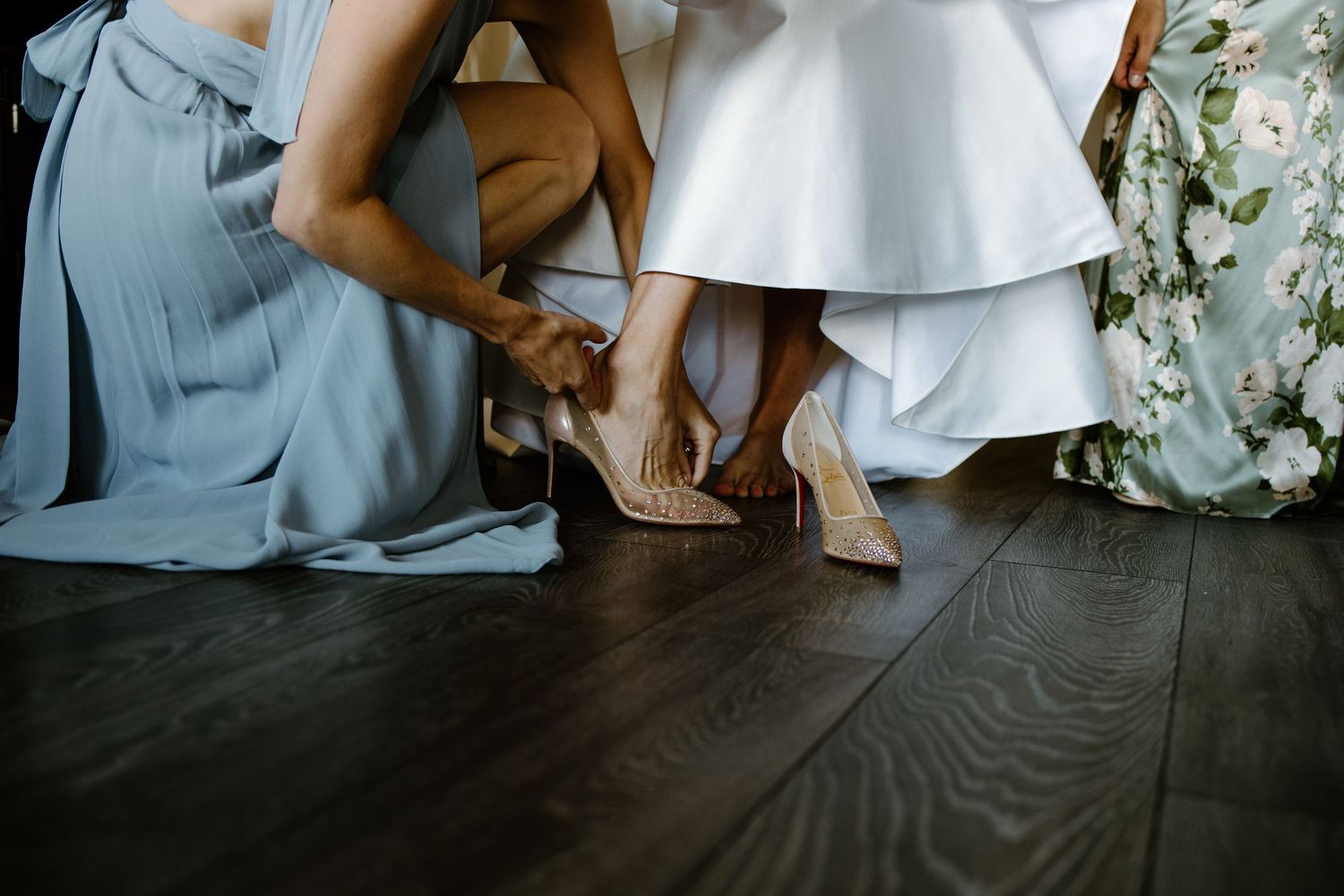 Bridesmaids helping bride put on elegant nude stiletto heels on dark hardwood floor before wedding ceremony.