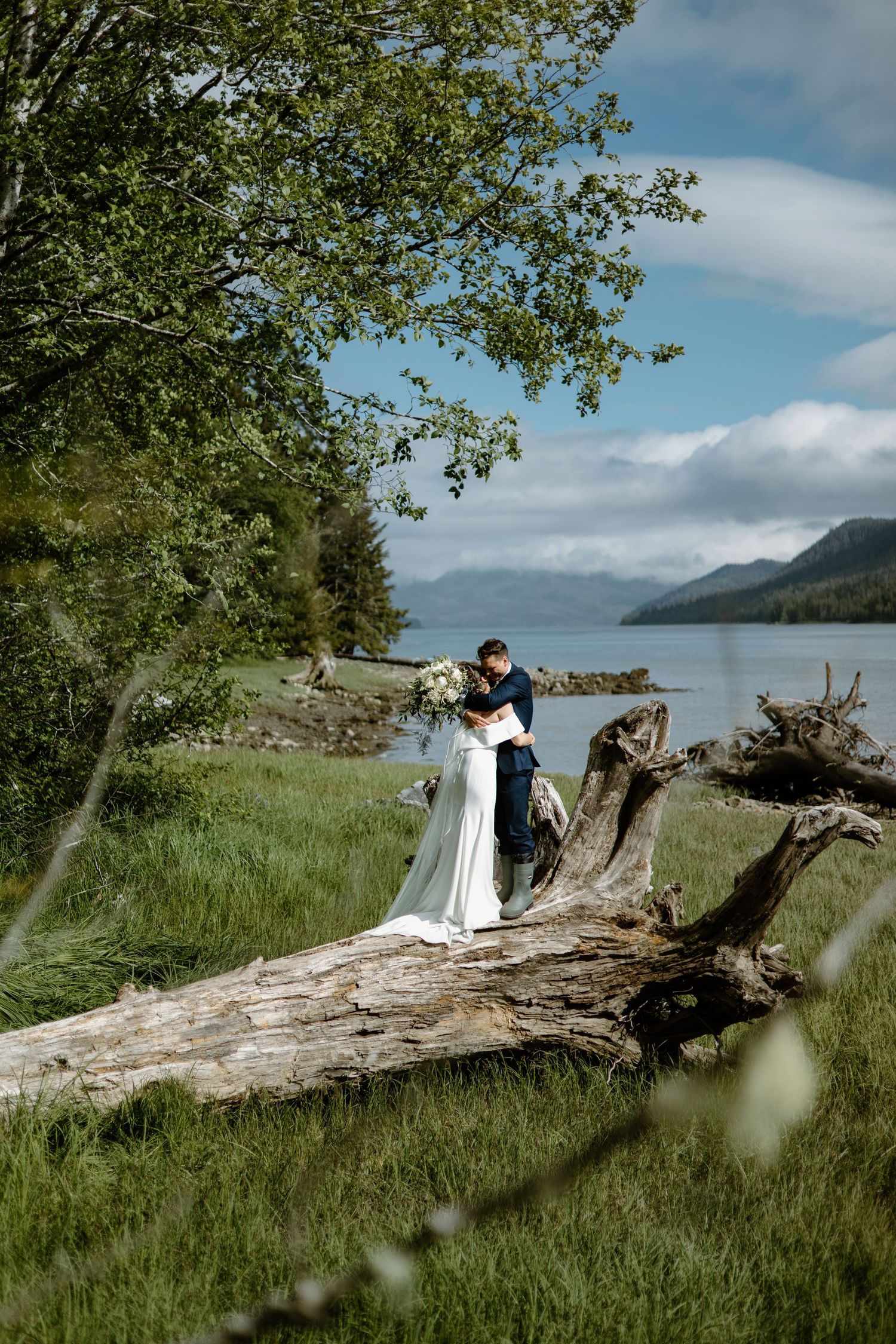 Bride and groom embrace on a fallen log by a scenic lake with mountains and lush greenery on their wedding day.
