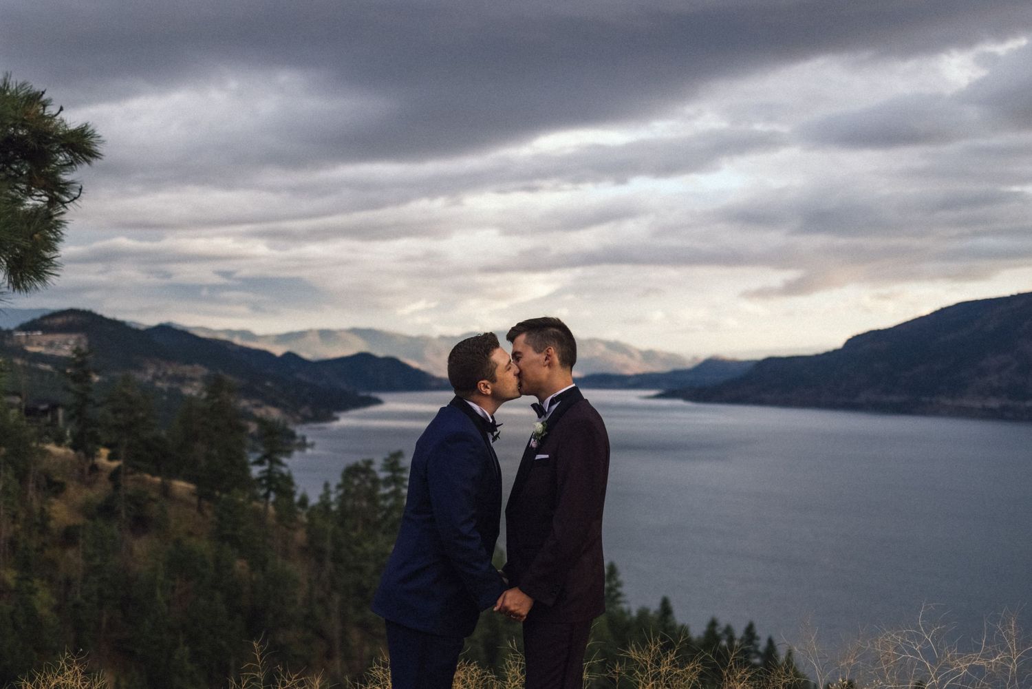 A couple shares a romantic moment overlooking a scenic mountain lake with dramatic cloudy skies above.