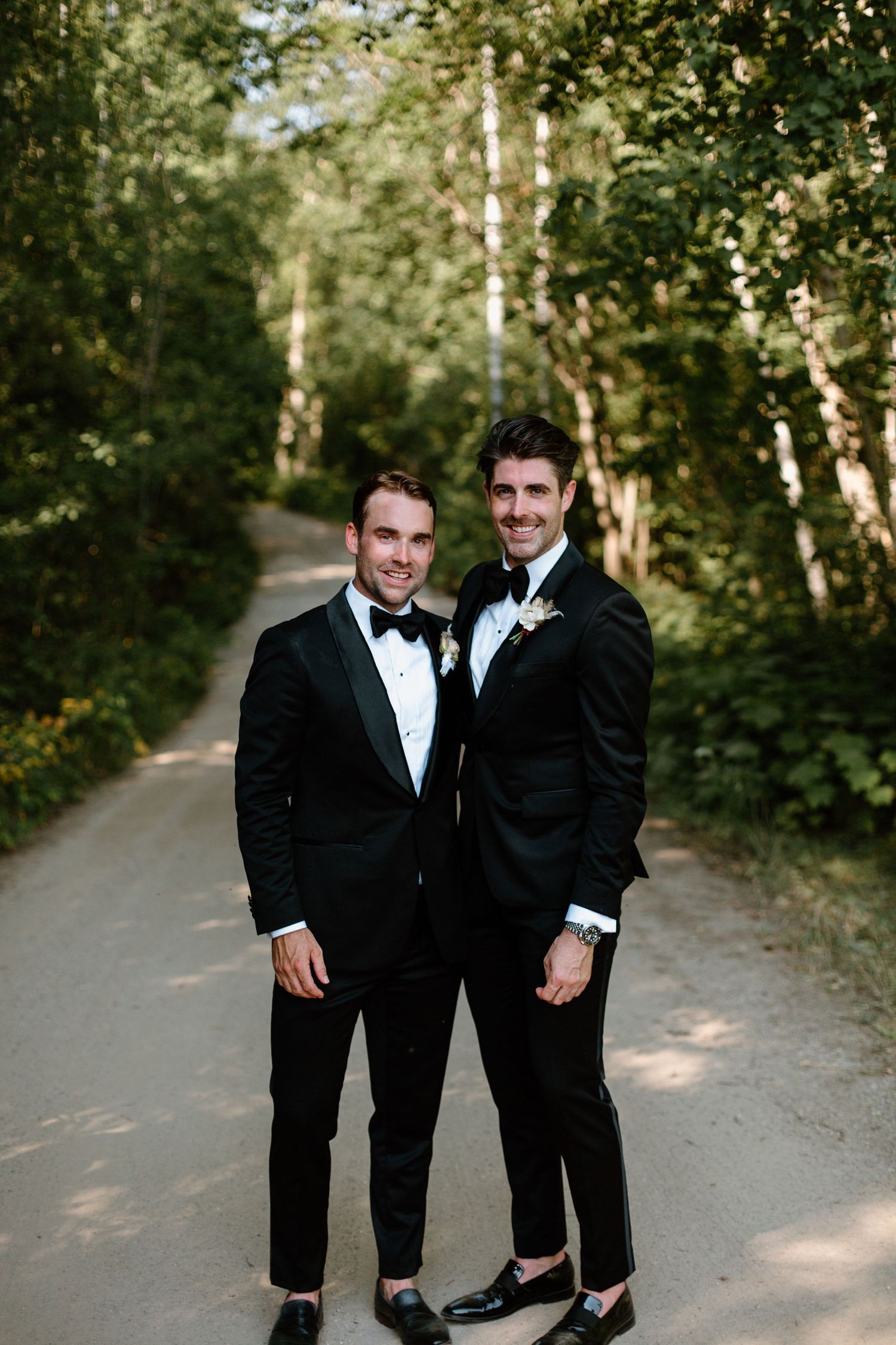 A couple in formal black tuxedos share candid moments together on a tree-lined path during a wedding photoshoot.