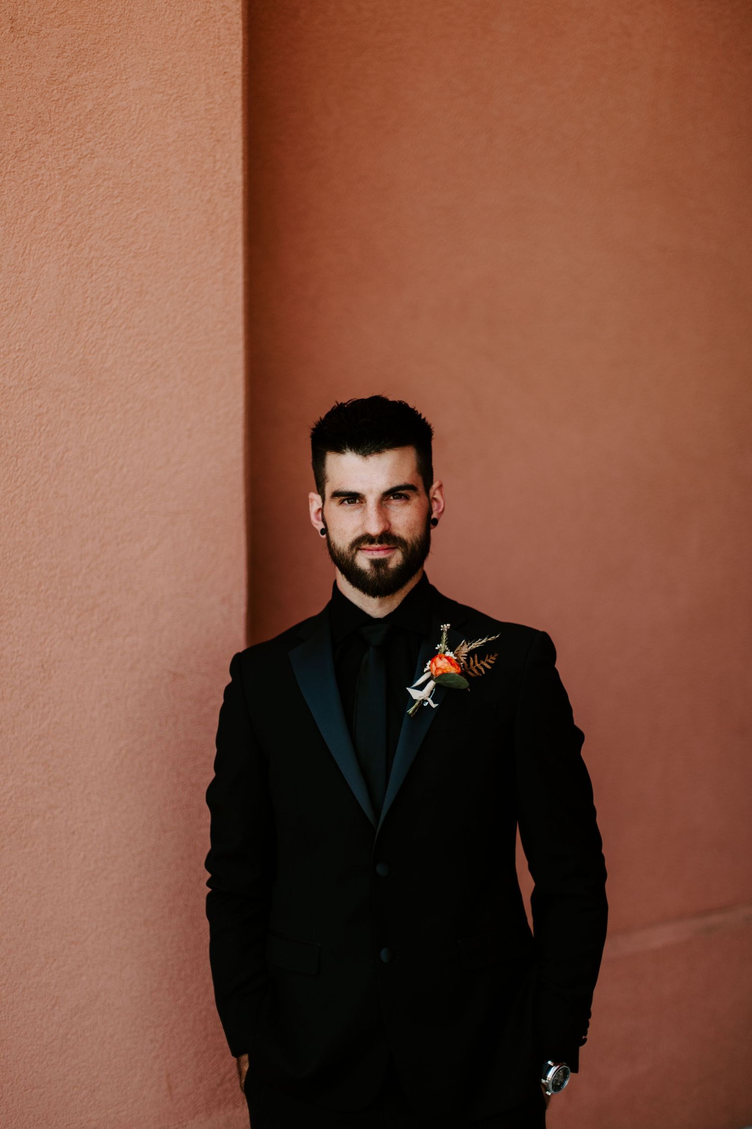 A person in formal black attire poses against a terracotta colored wall with dramatic lighting.