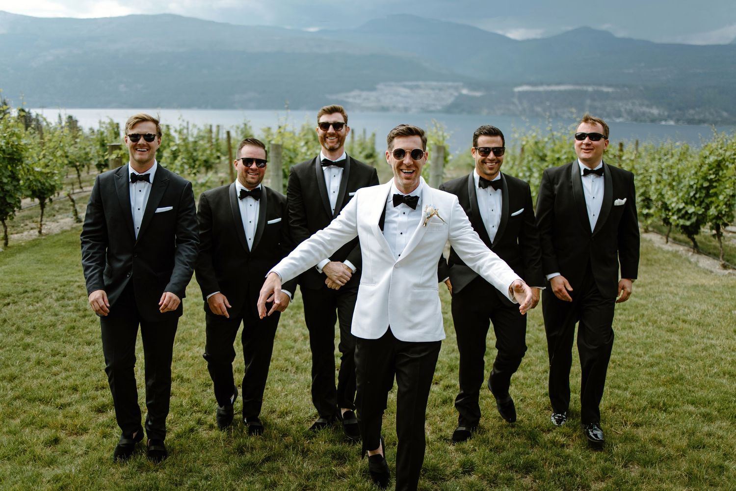 Groom in white tuxedo jacket leads groomsmen in black suits through vineyard with mountain lake backdrop.