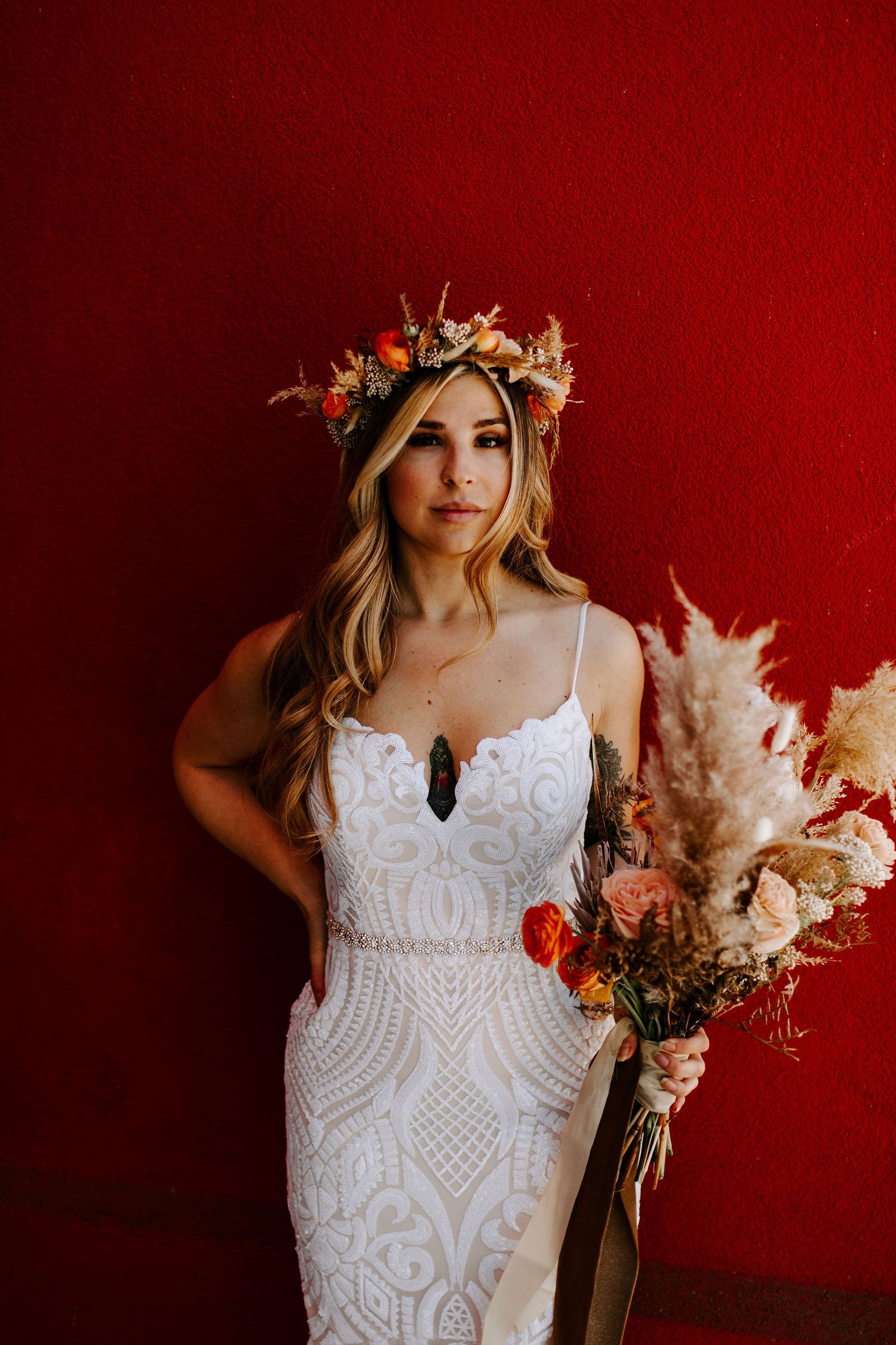 A person in a white lace dress holds dried flowers against a red wall wearing a floral crown.