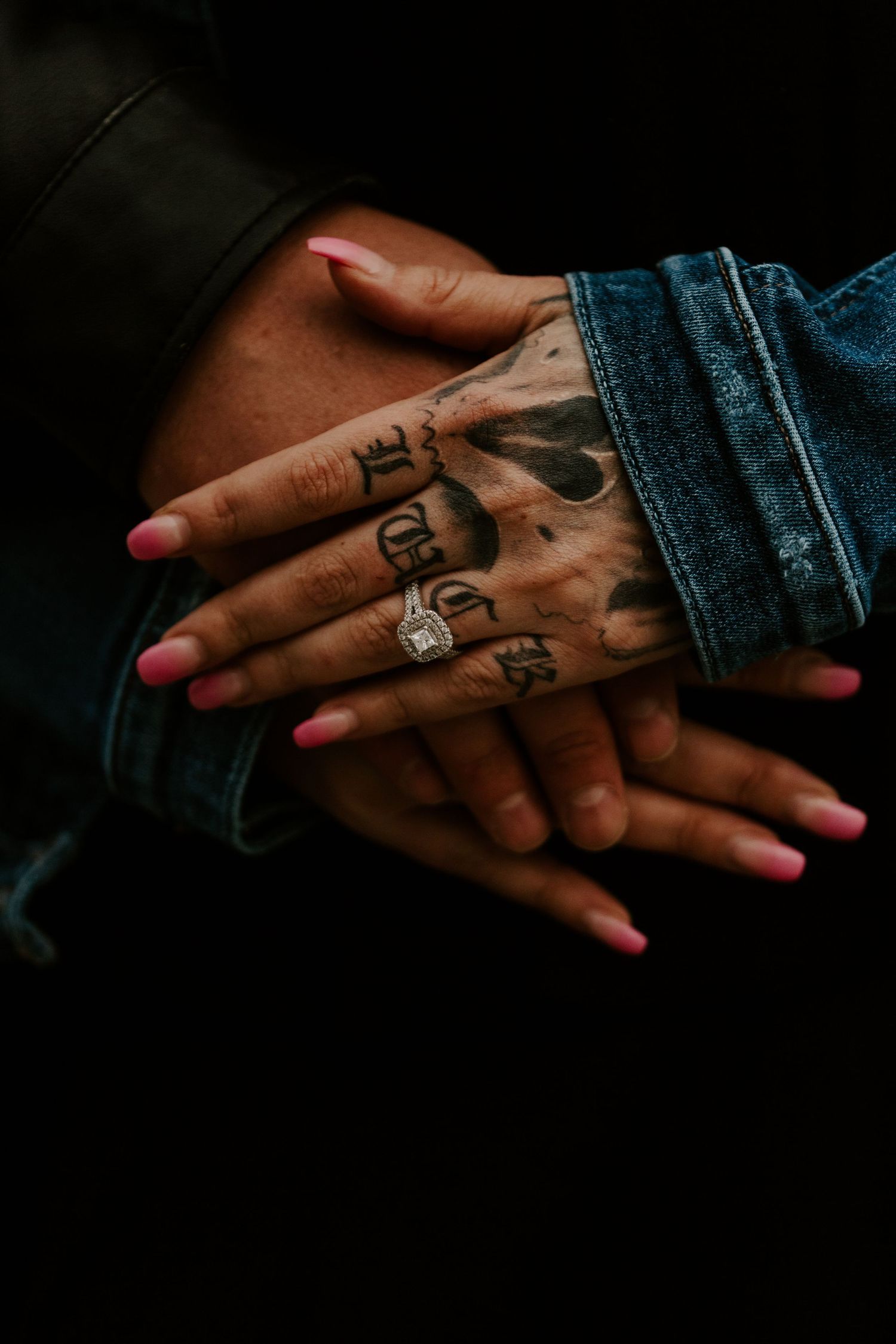 Tattooed hands with engagement ring shown against dark denim and leather background.