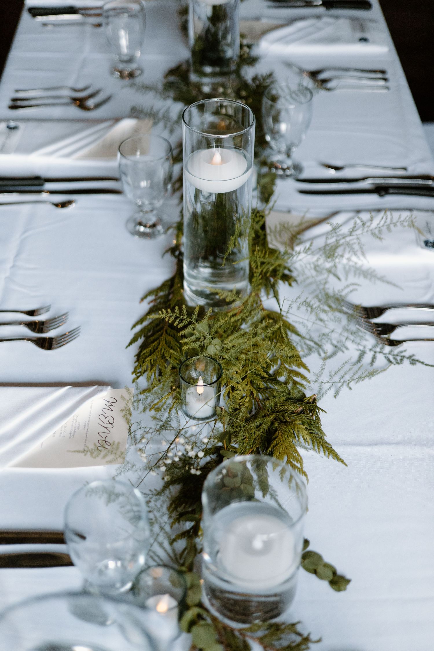Wedding table decorated with fern garland centerpiece and floating candles in glass cylinders.