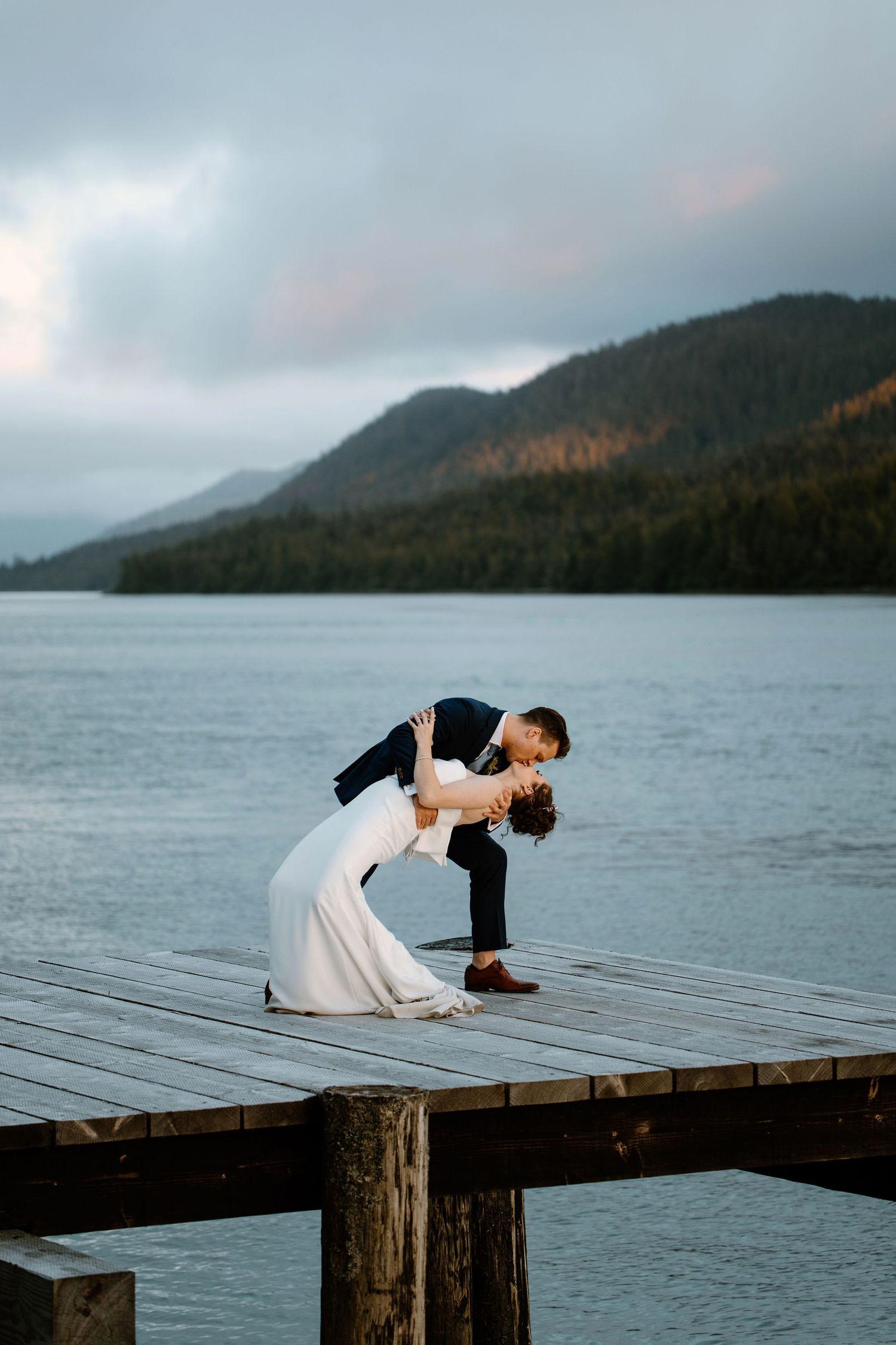 A couple performs graceful dance poses on a wooden dock overlooking a misty mountain lake at dusk.
