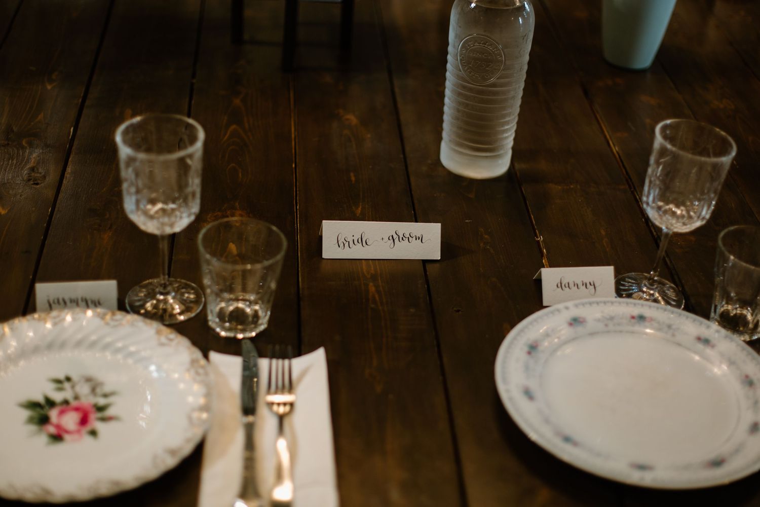 An elegant wedding place setting on a rustic wooden table with vintage plates and glassware.