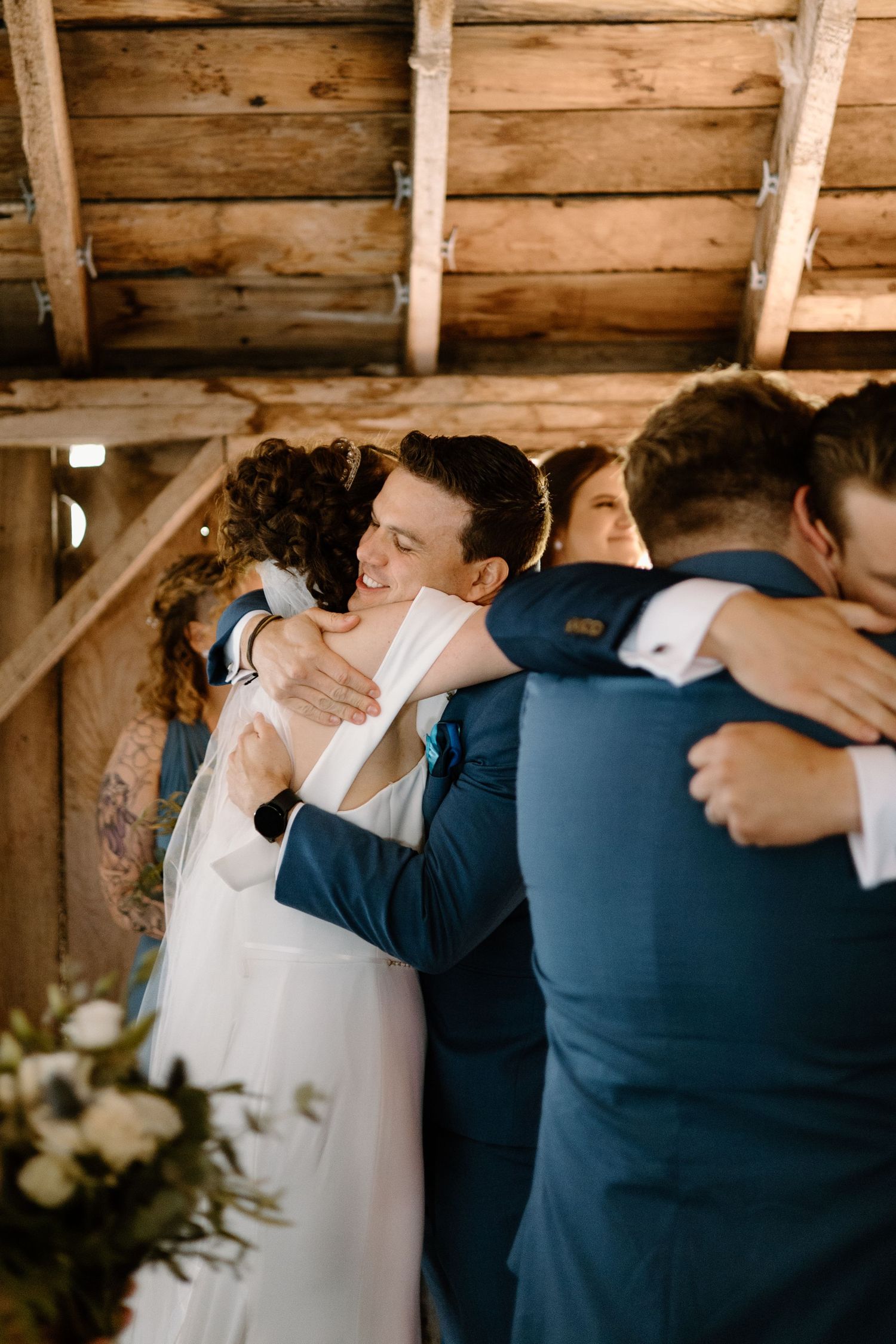 Emotional wedding party embrace during ceremony in wooden barn venue.