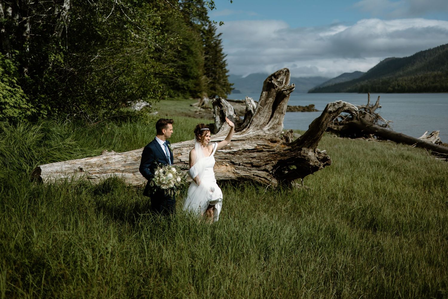 Bride and groom walk hand in hand through lush green grass beside a serene lake with a driftwood log backdrop.