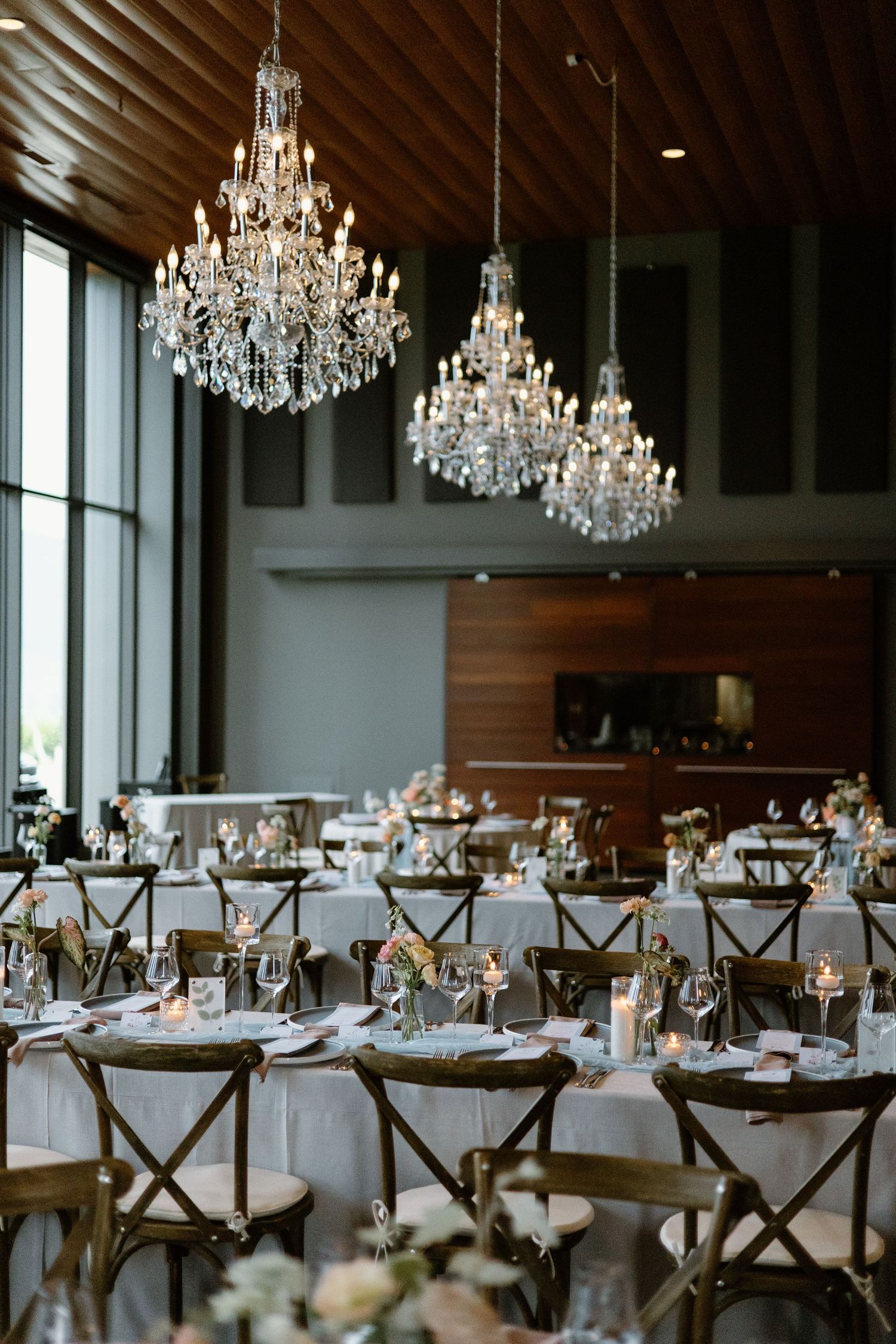 Elegant wedding reception hall with crystal chandeliers, round tables, white linens, and rustic cross-back chairs.