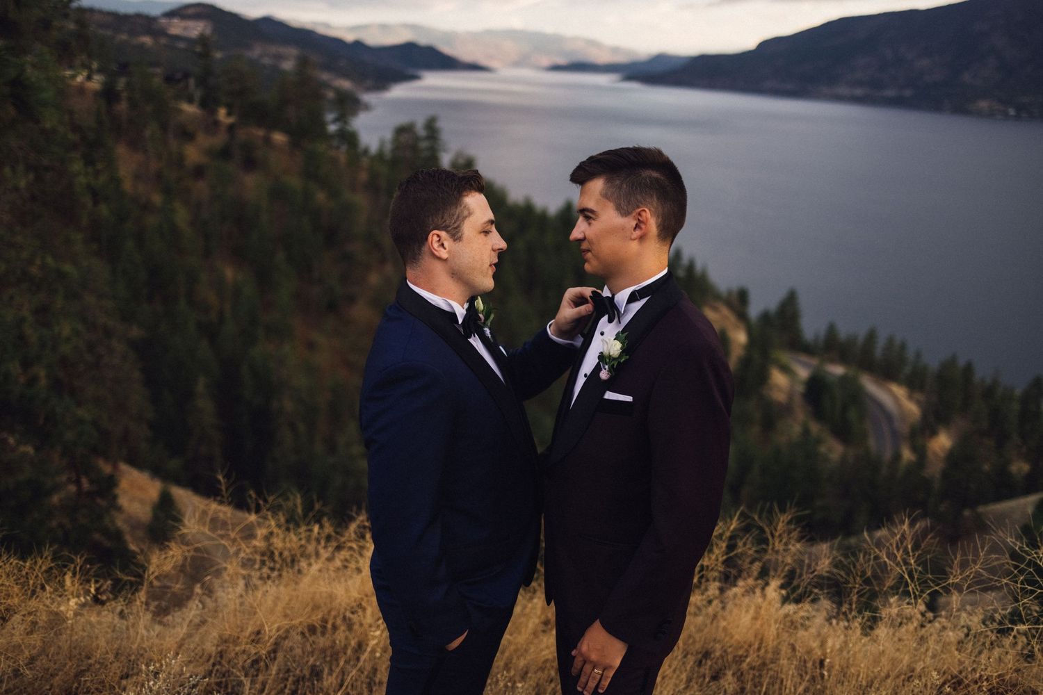 Two people in formal attire stand together on a scenic mountain overlook with a lake and hills in the background.