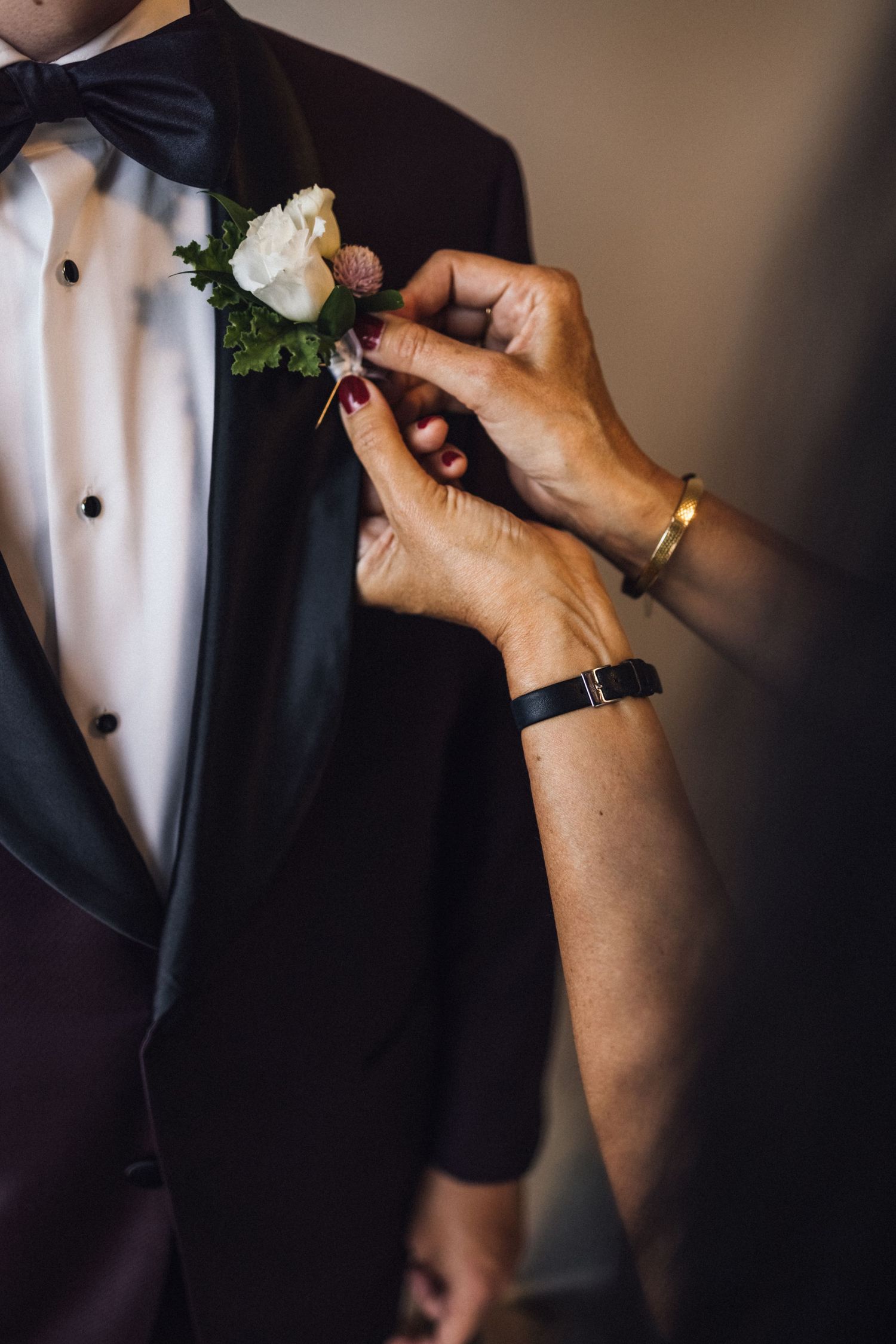 Hands pinning white rose boutonniere onto black tuxedo lapel.