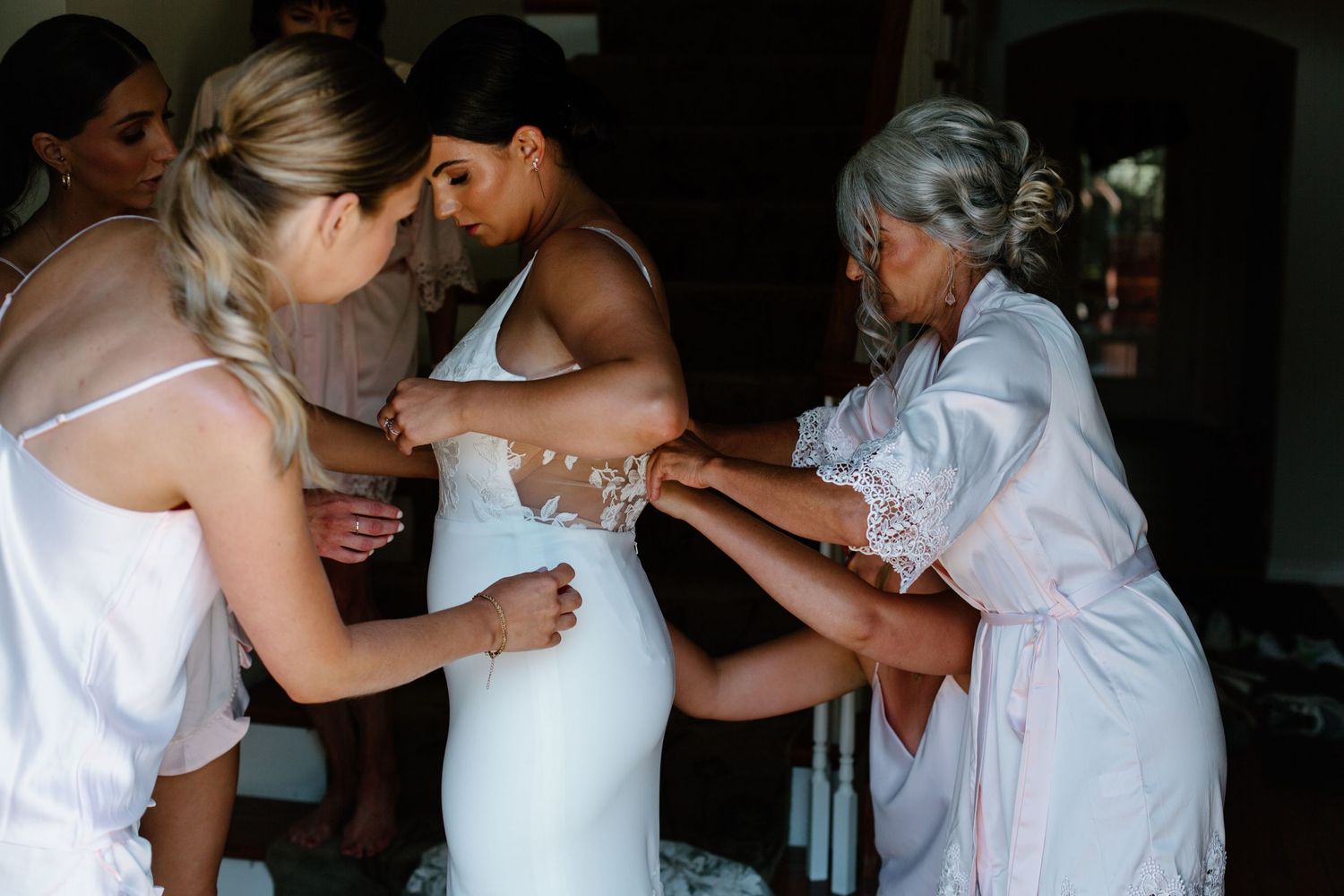 A touching moment during wedding preparations as helpers assist with the dress fitting.