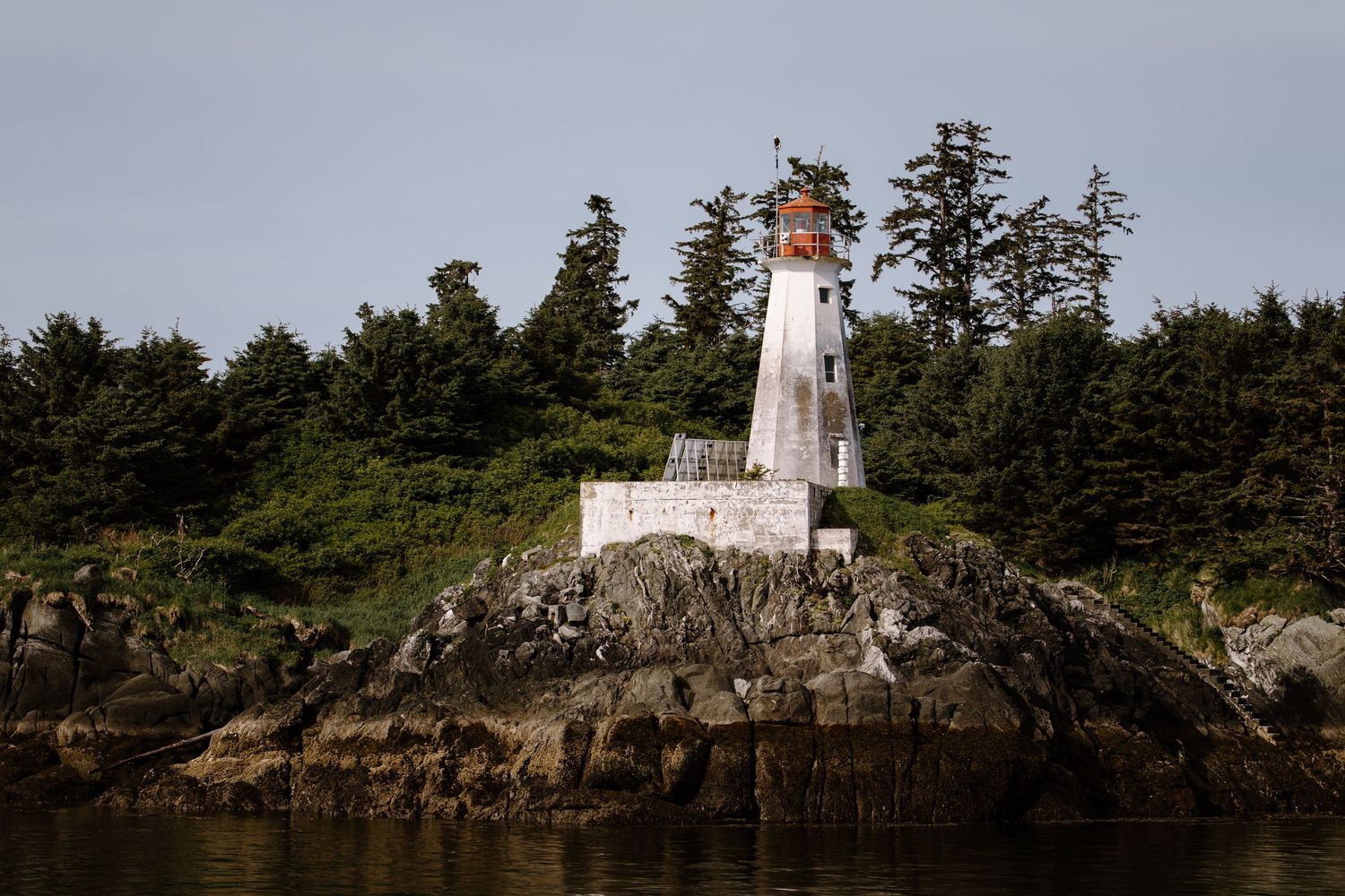 A white lighthouse perches on a rocky coastal cliff surrounded by evergreen trees.