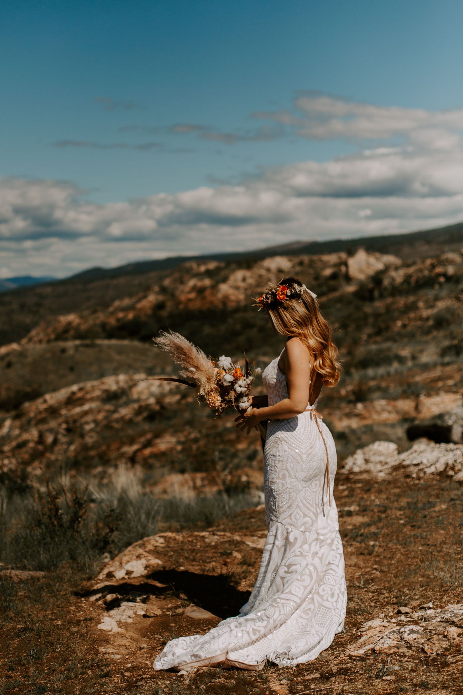 Person in white lace dress sitting on rocky terrain against a blue sky with mountains in the distance.