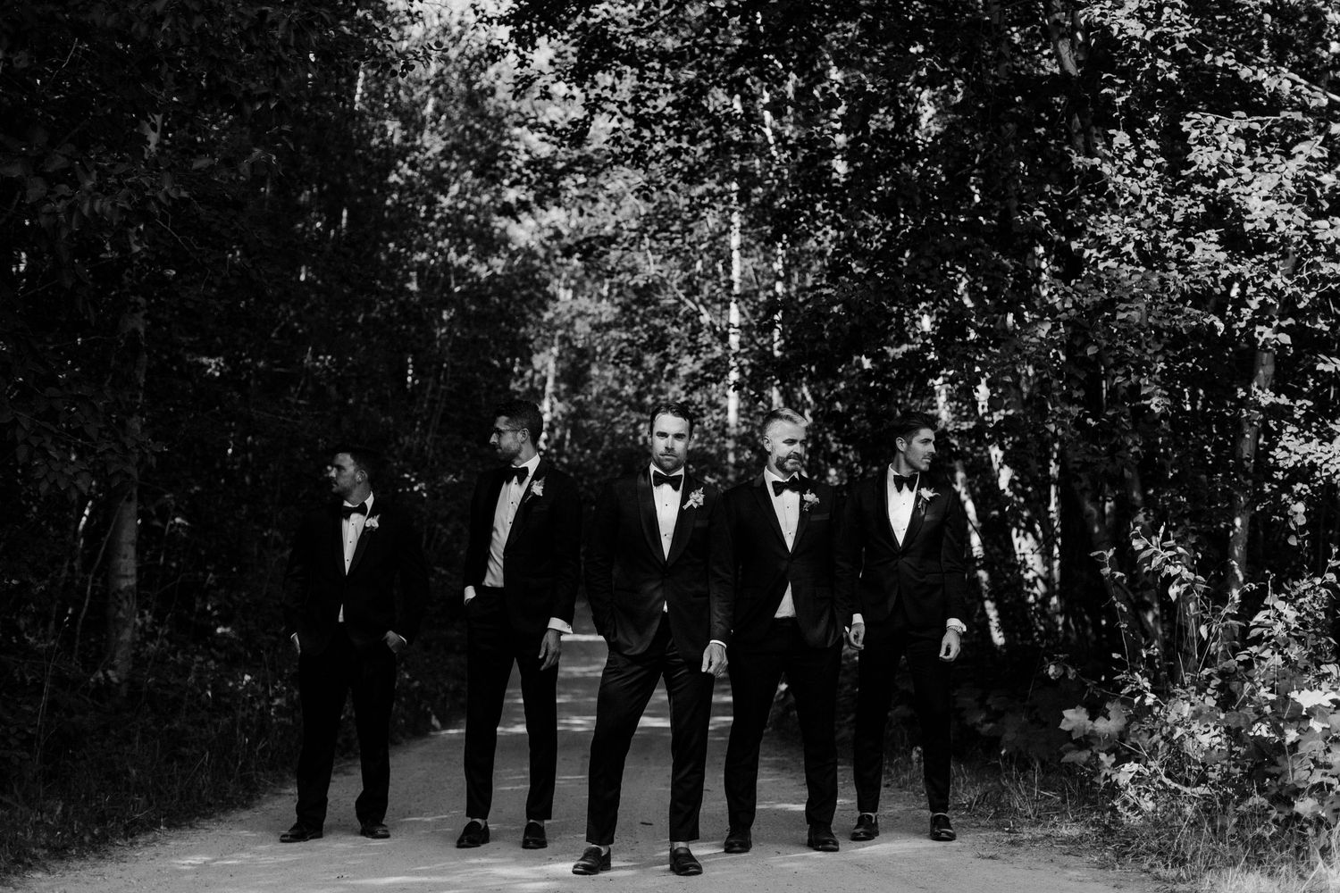 A black and white photo of five groomsmen in tuxedos standing on a wooded path.