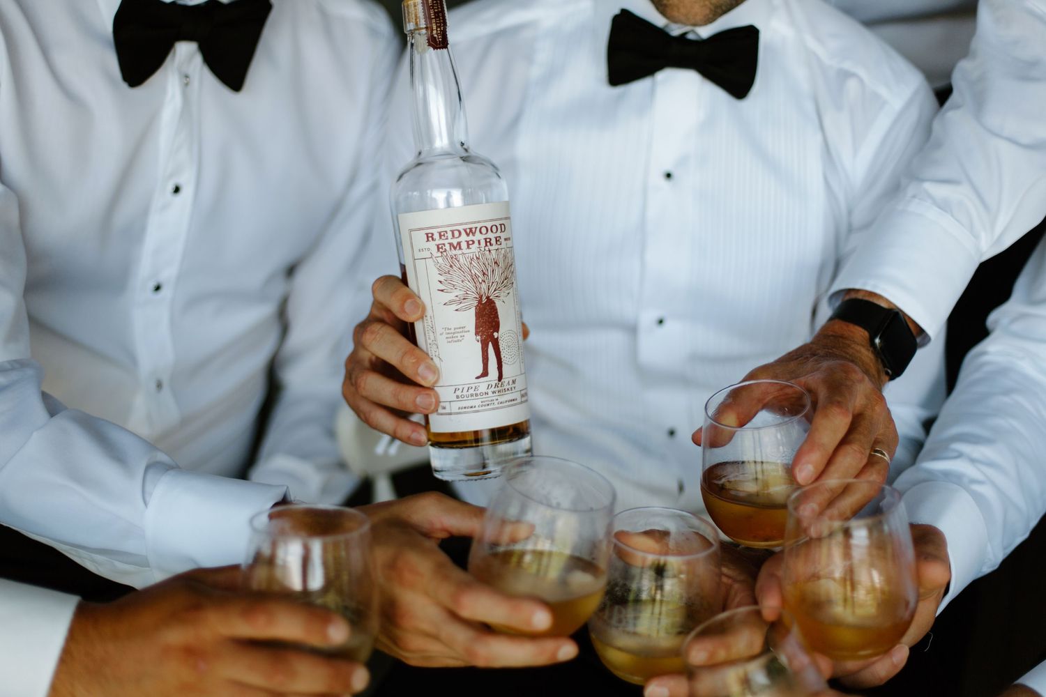 A person in a white dress shirt and bow tie pours a cocktail into a glass filled with ice.