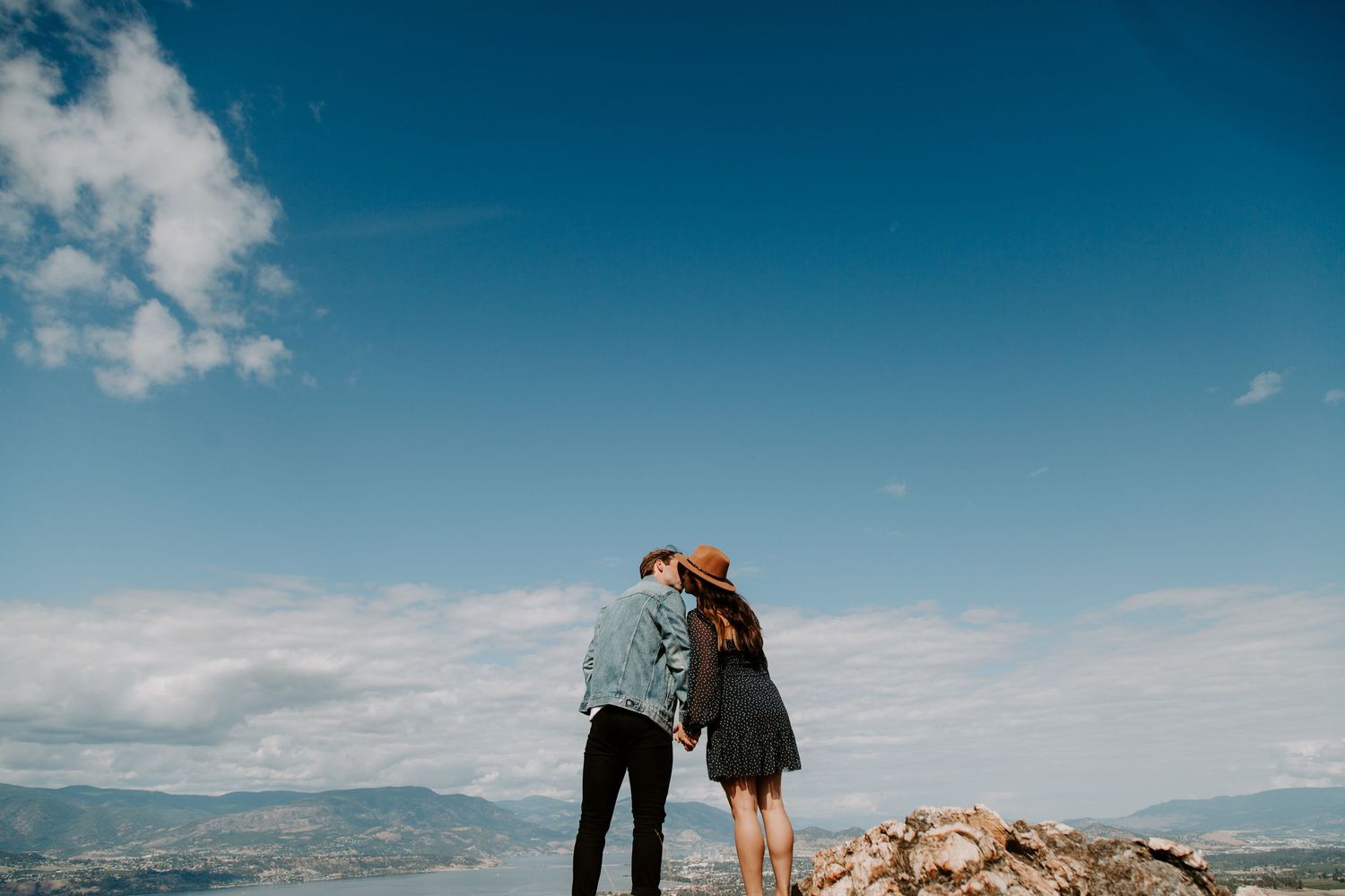 A couple shares an intimate moment while standing on a mountain summit against blue skies.