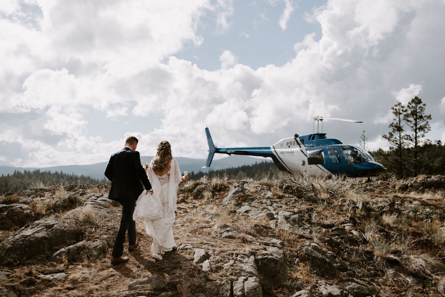 Newlyweds walking near helicopter on mountain terrain with cloudy sky backdrop.